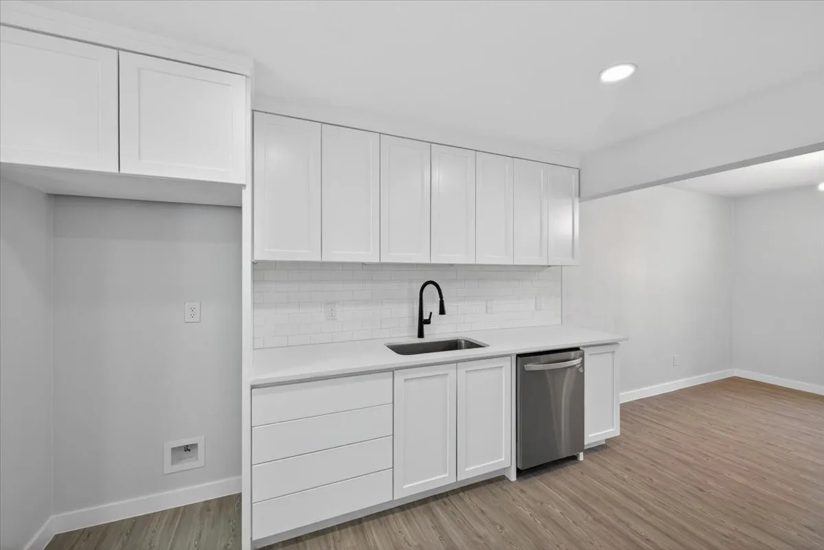 Contemporary kitchen area featuring white shaker-style cabinetry, a stainless steel dishwasher, a black gooseneck faucet, white subway tile backsplash, and wood-finish flooring