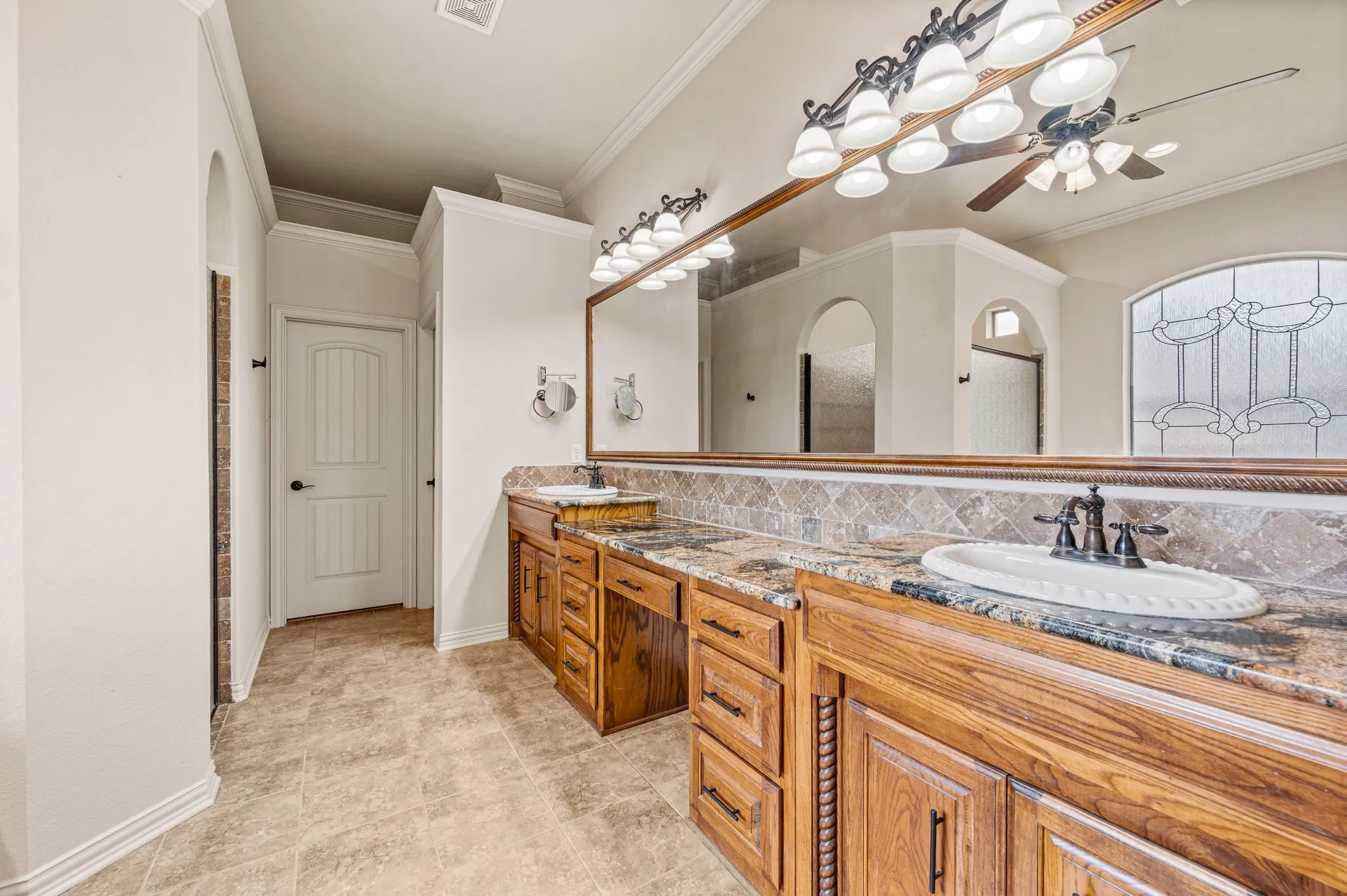 The Primary bath has travertine flooring, custom dual sink cabinetry with vanity seating, framed mirror and wrought iron light fixtures. Door ahead leads into a large wardrobe closet.