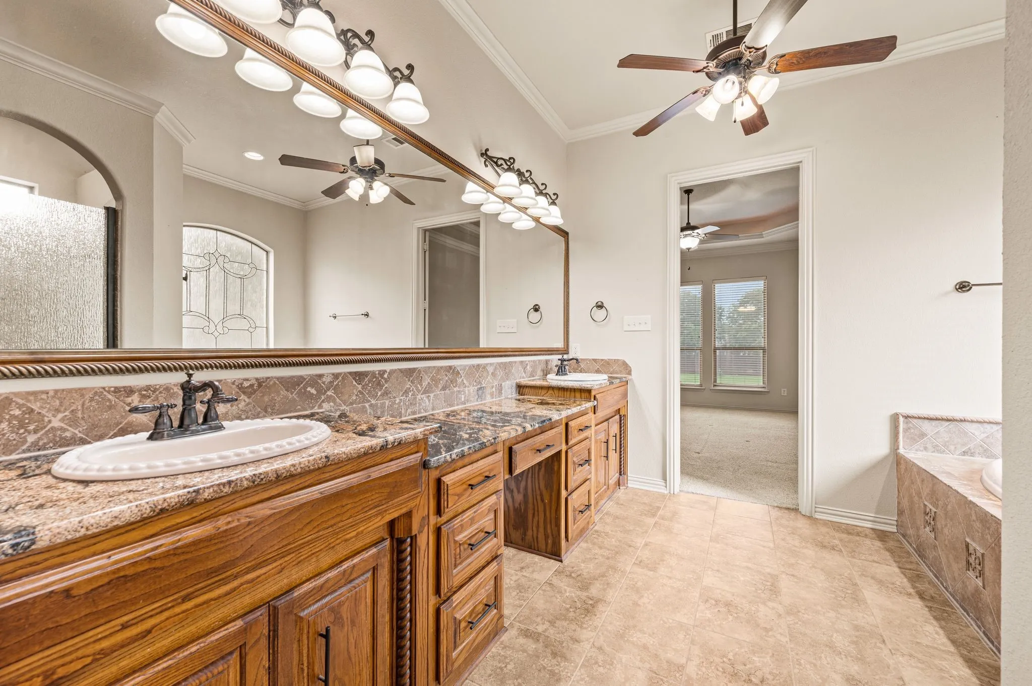 Another view of the Primary bath. Granite topped spacious vanity with decorative oval sinks and vanity space seating. Bronze fixtures and hardware, a ceiling fan and crown molding provide a classic look.