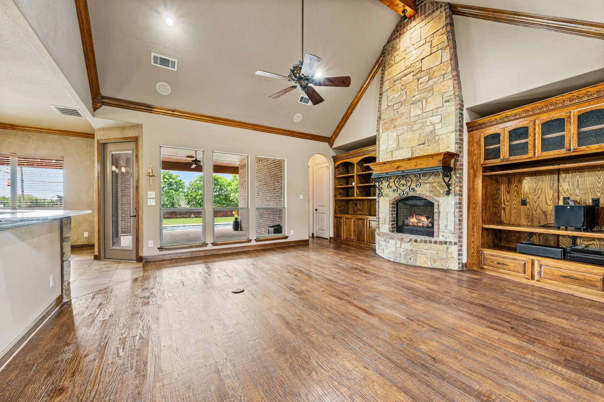A floor to ceiling stone, gas log fireplace with a wood and iron mantle and hearth is flanked by custom wood built in cabinetry. Crown moldings and an exposed wood beam provide architectural details to the vaulted ceiling. Hand scraped wood floors, surround sound, ceiling fan, a floor electrical outlet, and a wall of windows to the back patio and yard compliment this space.