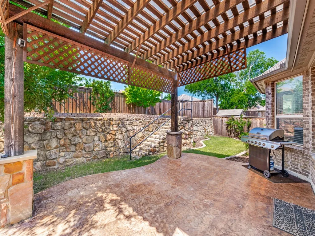 Expansive rear patio featuring a wood pergola with lattice detailing, stamped concrete flooring, a stone retaining wall, and a staircase with metal railings