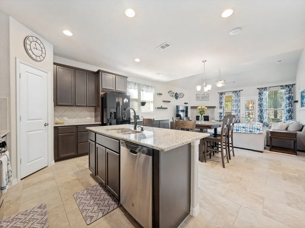 Open-concept kitchen featuring an island with a built-in sink and dishwasher, dark wood-finish cabinetry, granite countertops, and tile flooring