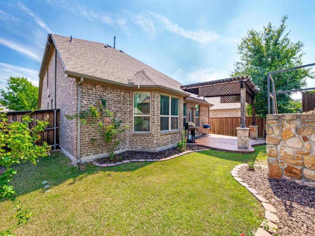 Expansive backyard featuring a brick exterior, a covered patio with a lattice pergola, a stone-faced retaining wall, manicured lawn, and perimeter landscaping
