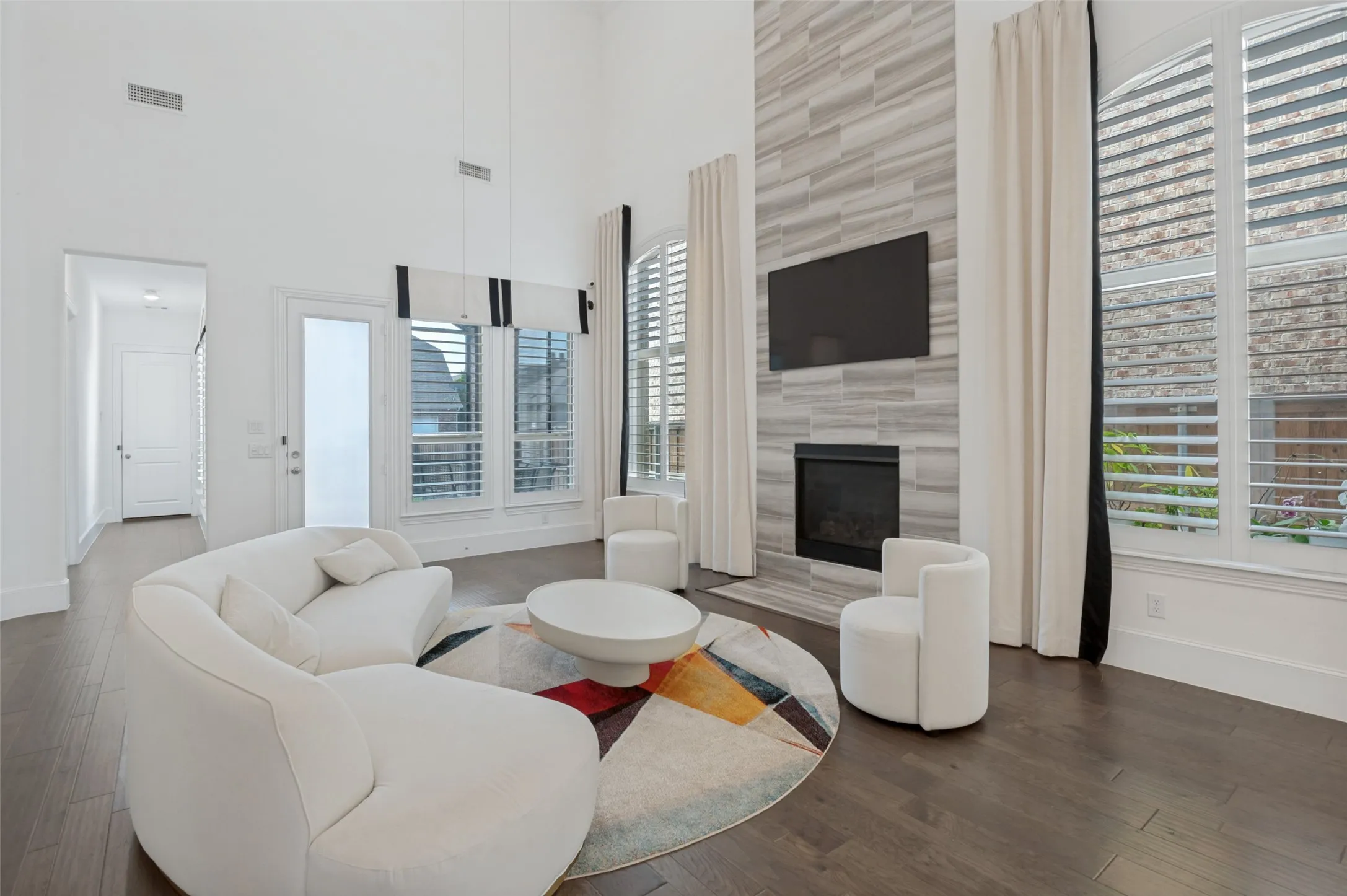 Spacious living area featuring a floor-to-ceiling tiled fireplace surround, high ceilings, dark wood-finish flooring, and large windows with plantation shutters