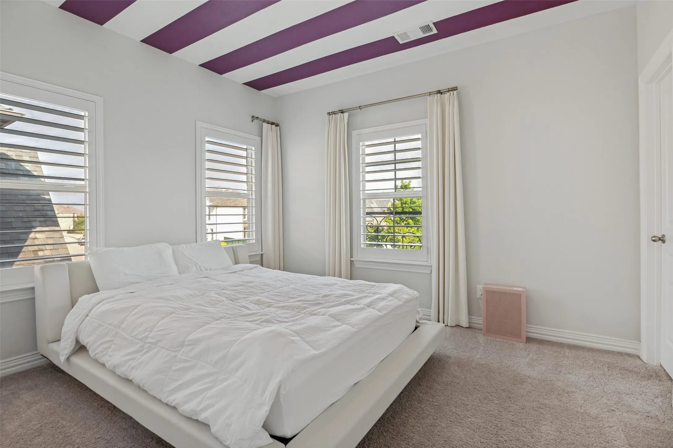 Bedroom featuring a distinctive ceiling with purple and white stripes, two windows with white plantation shutters, and light-toned carpeting