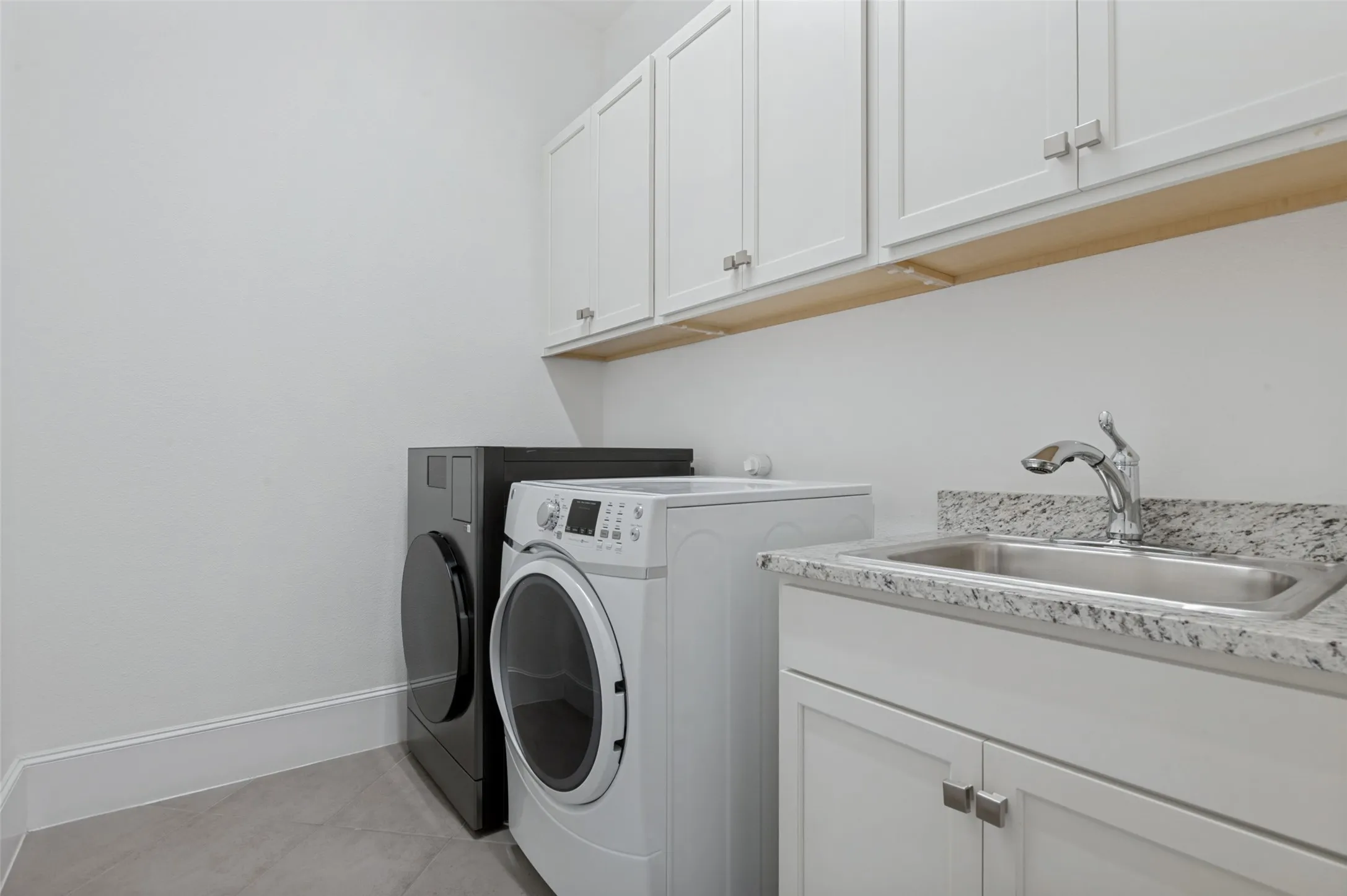 Dedicated laundry area featuring white shaker-style cabinetry, granite-finish countertops, and a stainless steel utility sink with a chrome faucet