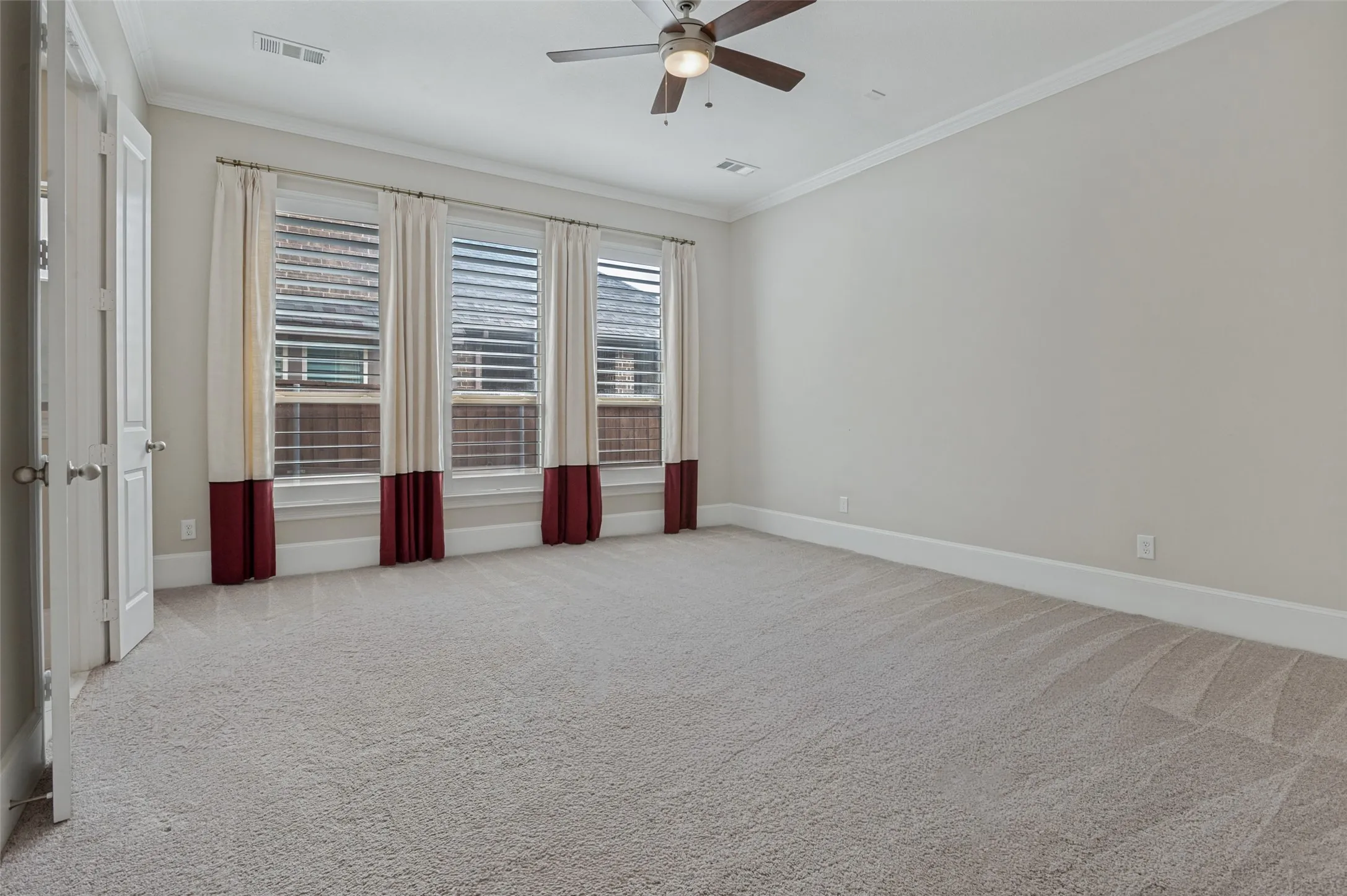 Spacious room featuring light-colored carpeting, three large windows with blinds and decorative drapes, and a ceiling fan with integrated lighting