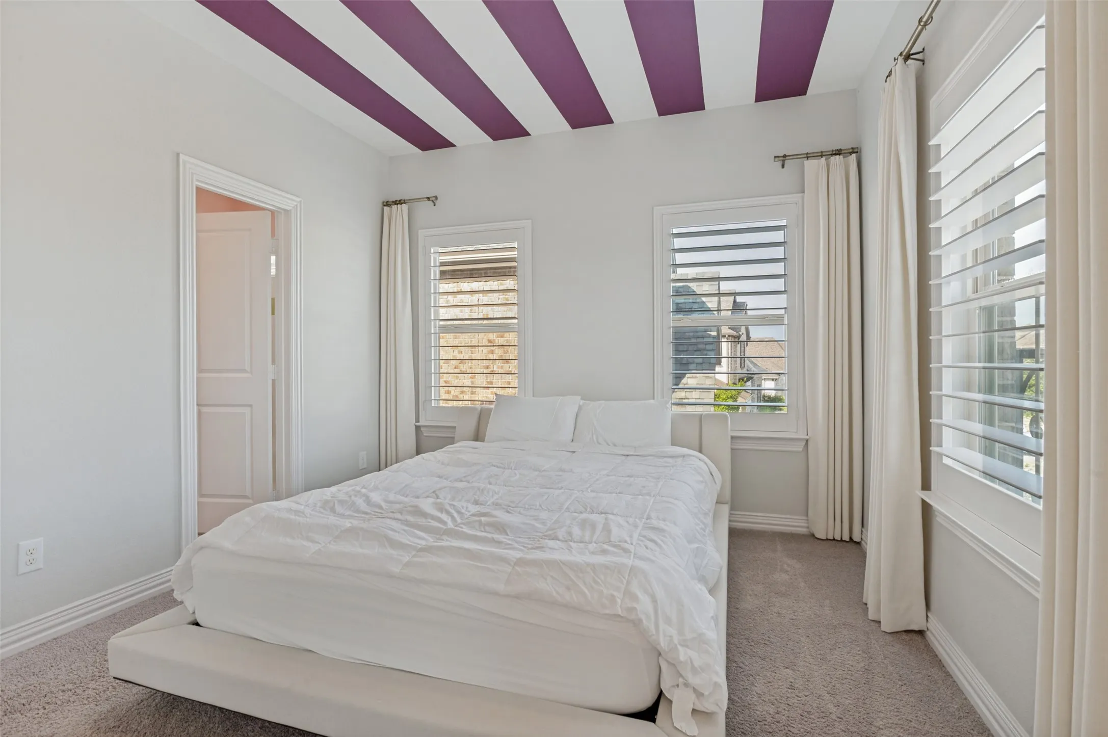 Bedroom featuring a tray ceiling with accent stripes, two windows with plantation shutters, full-length drapes, and neutral carpeting