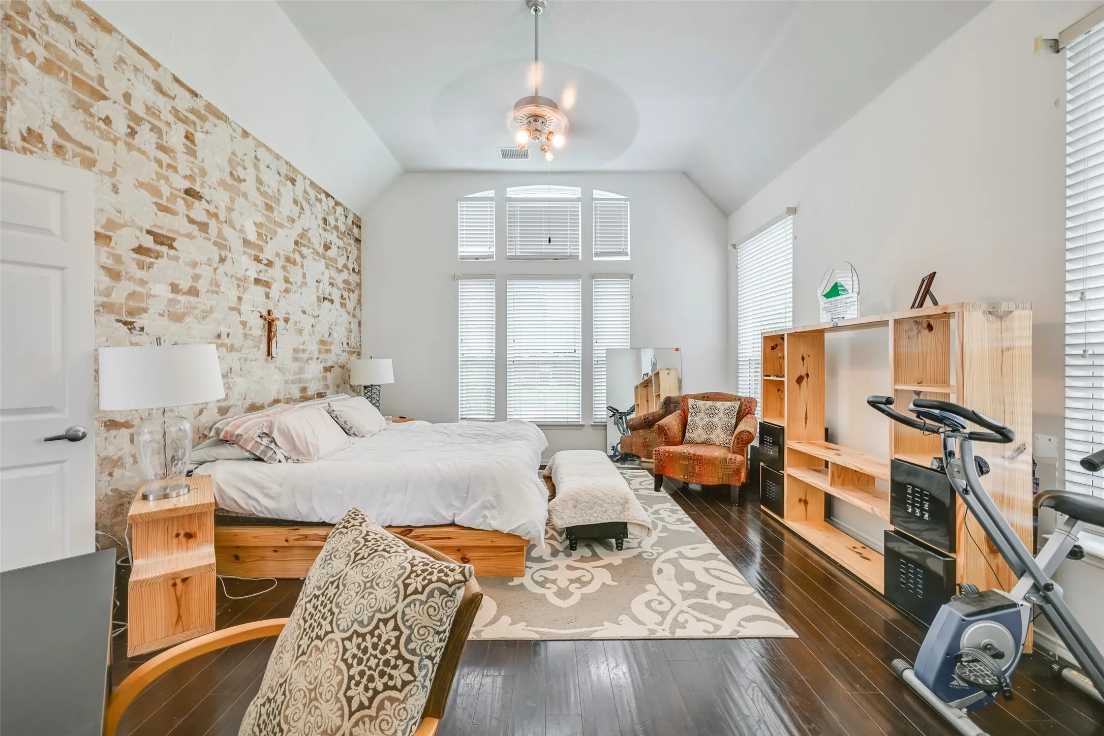 Vaulted ceiling bedroom featuring a whitewashed brick accent wall, dark wood-finish flooring, and expansive windows with integrated blinds