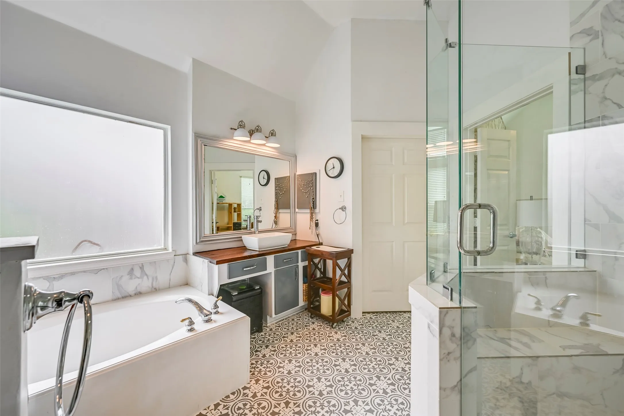 Spacious bathroom featuring a glass-enclosed shower, built-in soaking tub with marble-look surround, patterned floor tiling, and a vanity with a vessel sink