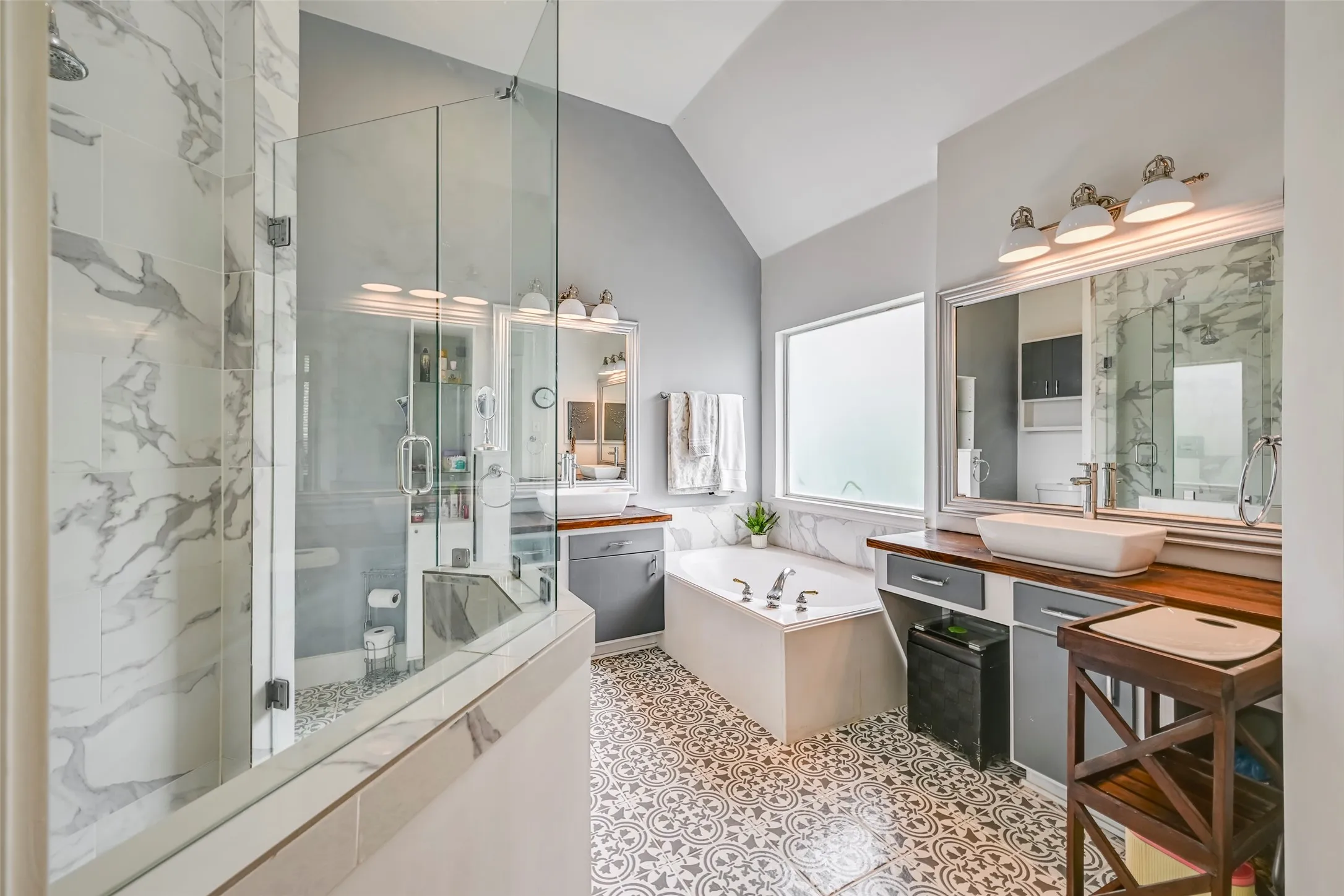 Bathroom featuring a walk-in shower with glass enclosure and marble-look tile, a built-in soaking tub, dual vanities with vessel sinks and wood-finish countertops, and patterned floor tiling