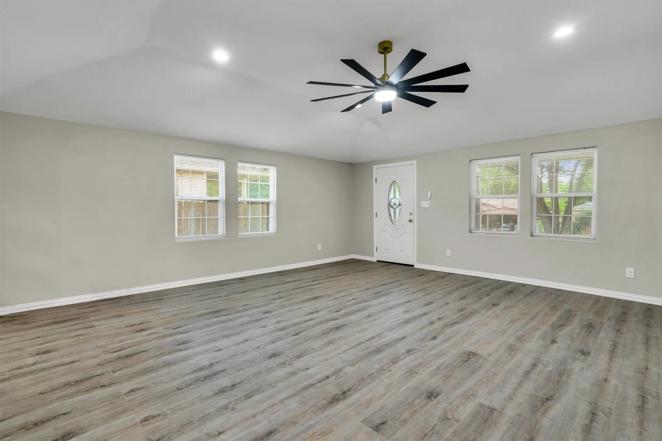 Spacious room featuring wood-finish flooring, a modern ceiling fan with lighting, recessed ceiling lights, multiple windows with white trim, and a white entry door with decorative glass