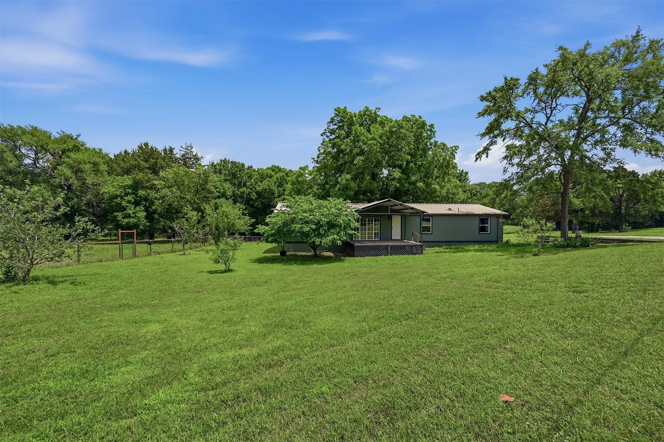 Expansive green lawn surrounding a light blue single-story residence with a covered porch and white columns, framed by mature trees
