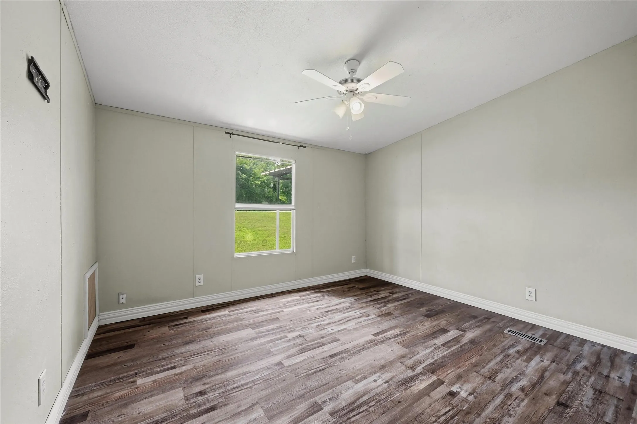 Spacious room featuring wood-finish flooring, light-toned walls, a window with exterior views, white baseboards, and a ceiling fan with integrated lighting