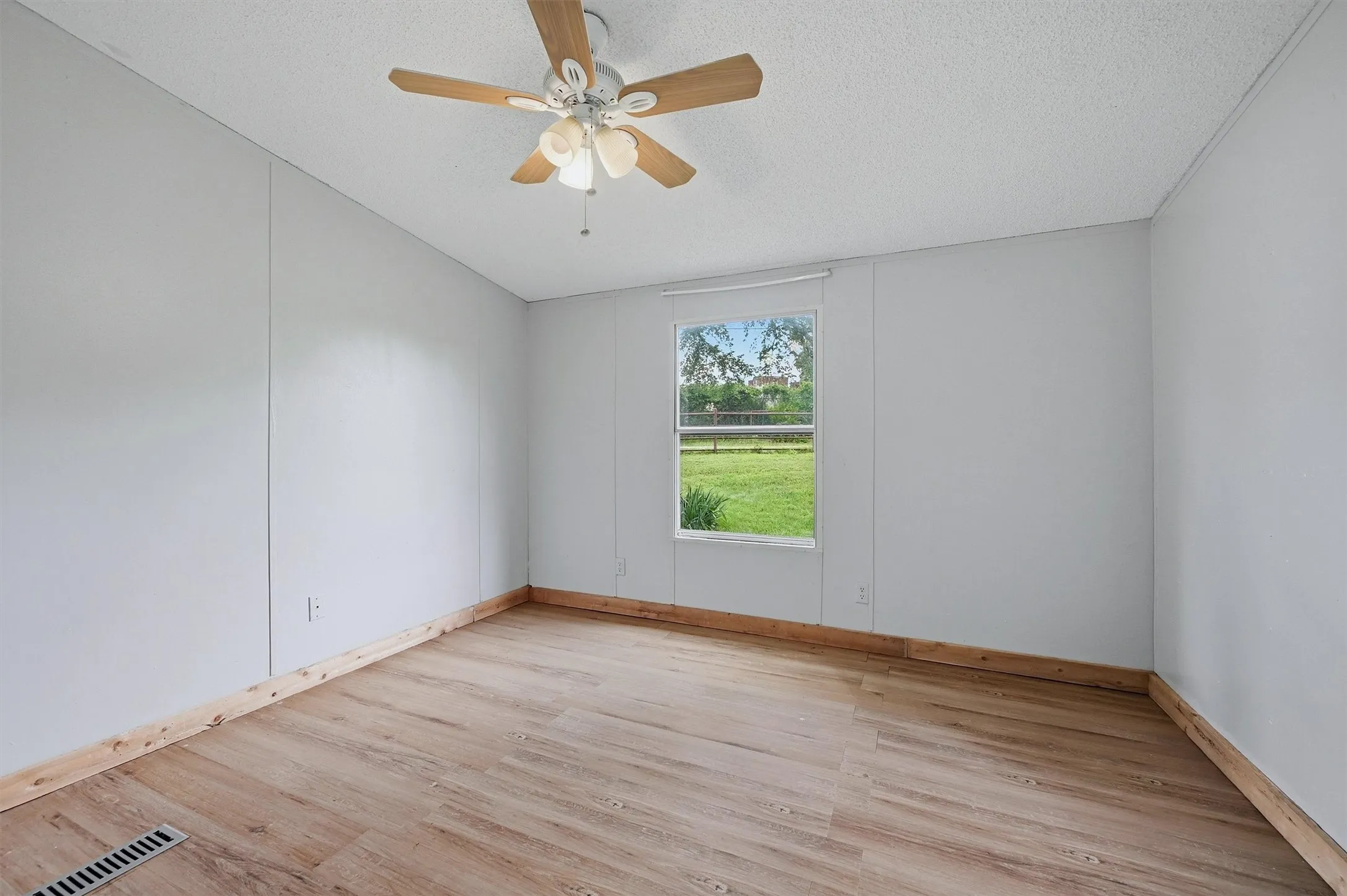 Spacious room featuring light wood-finish flooring, light gray wall panels, a white ceiling with a ceiling fan, and a window offering exterior scenery
