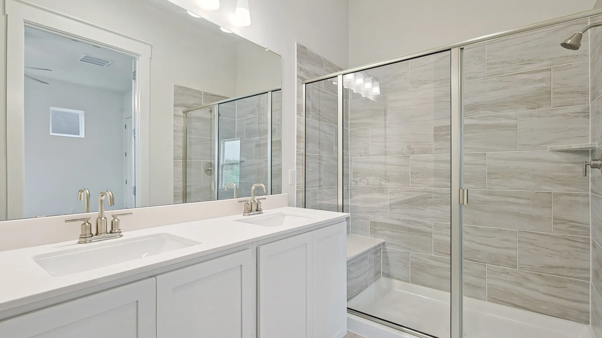 Modern bathroom featuring a dual vanity with white cabinetry and integrated sinks, brushed nickel fixtures, a full-width mirror, and a tiled walk-in shower with a glass enclosure and built-in bench