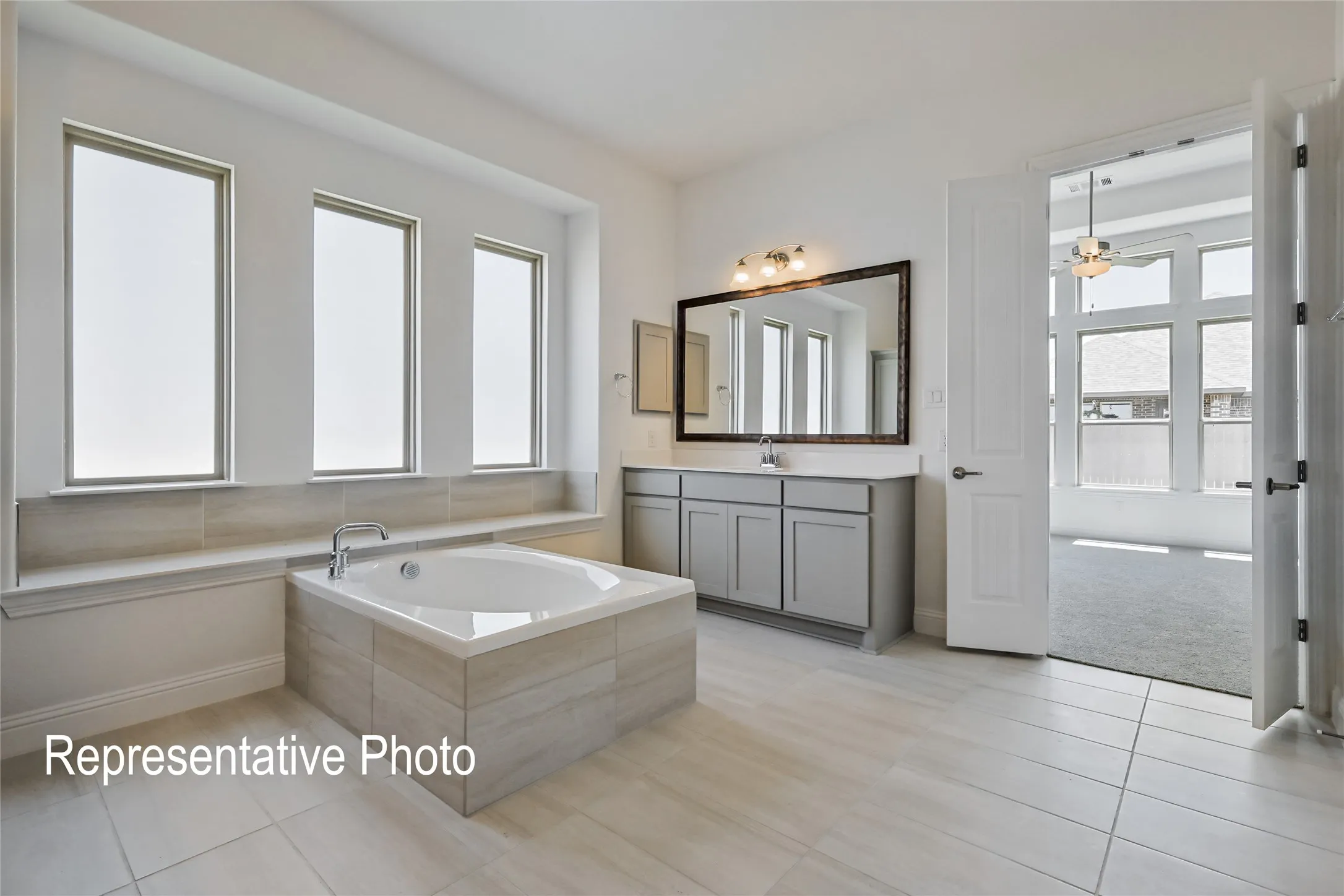 Spacious bathroom featuring a built-in soaking tub with tiled surround, three vertical windows, a dual-sink vanity with light-colored cabinetry, and large format tile flooring