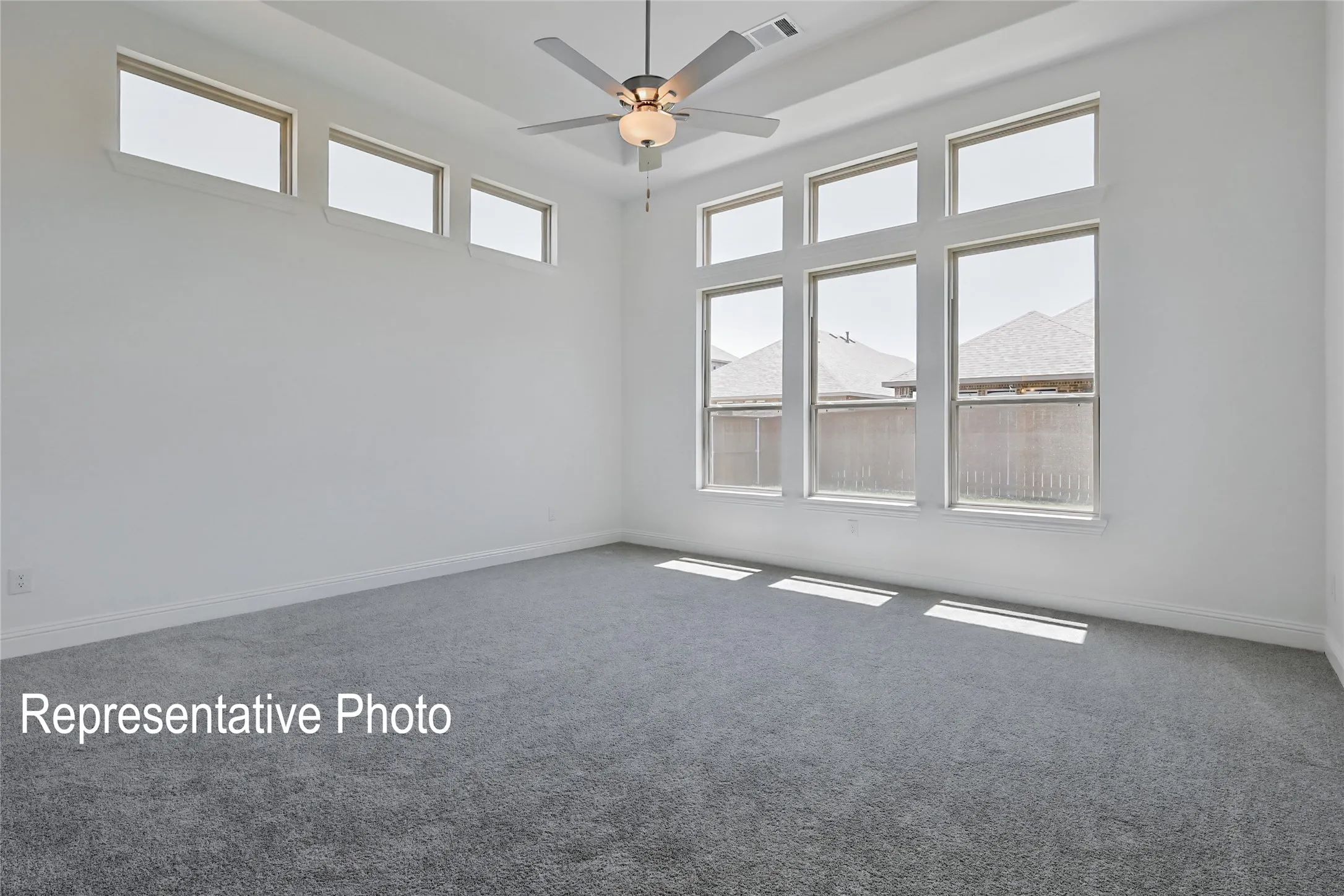 Spacious room featuring gray carpeting, white walls, multiple large windows, a ceiling fan with integrated lighting, and a high ceiling