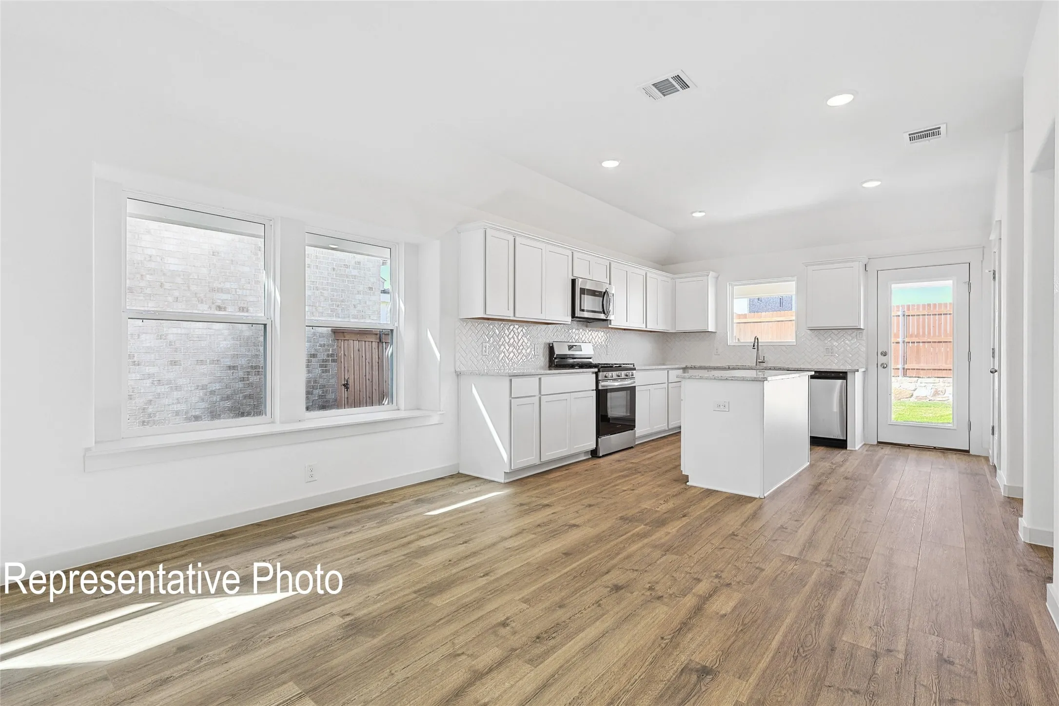 Open-concept living space featuring wood-finish flooring, a kitchen island with stone countertop, white shaker cabinetry, stainless steel appliances, and multiple windows