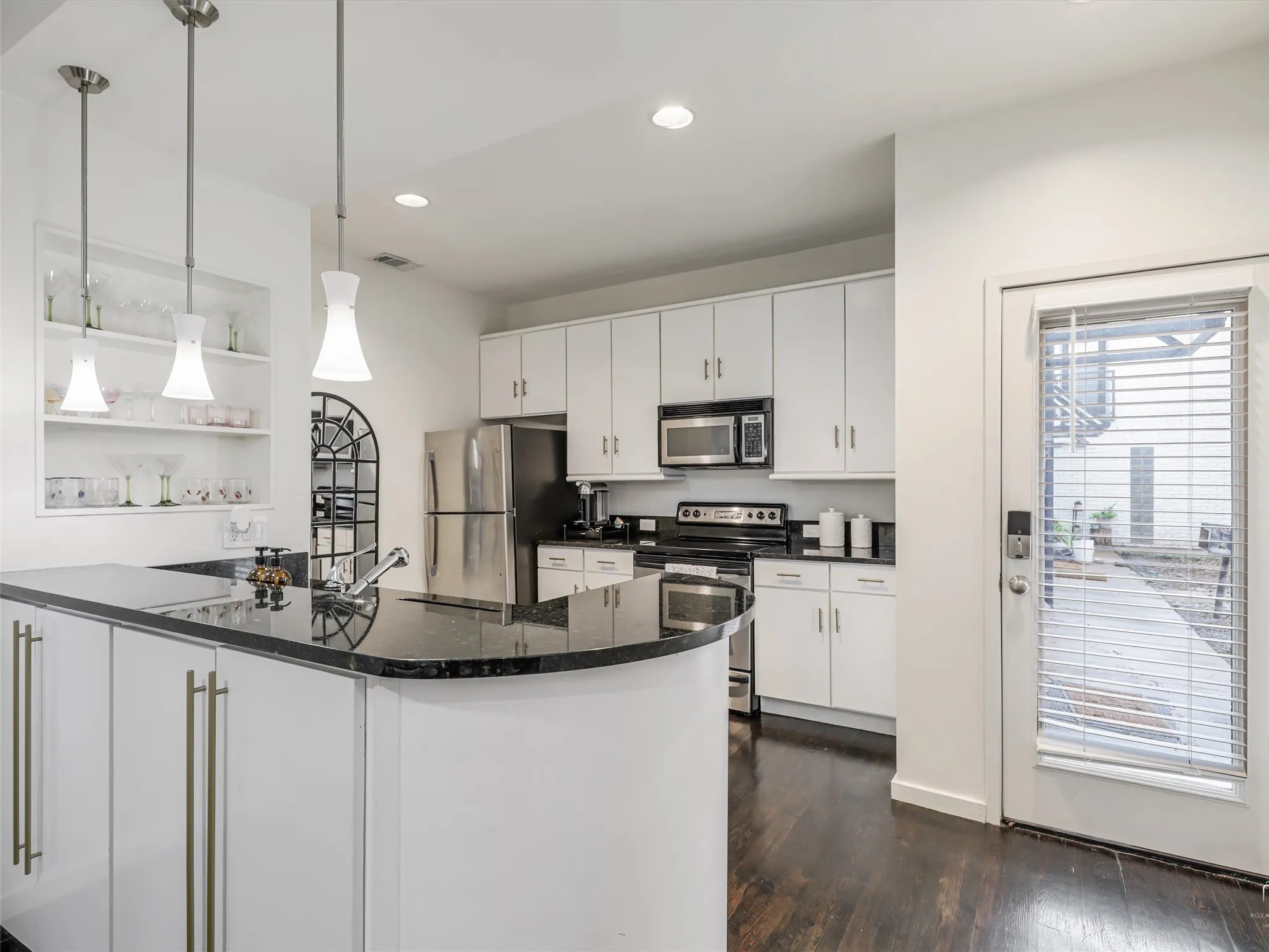Modern kitchen featuring an island with black countertop, stainless steel sink, white cabinetry with brushed nickel hardware, built-in shelving, and dark wood-finish flooring