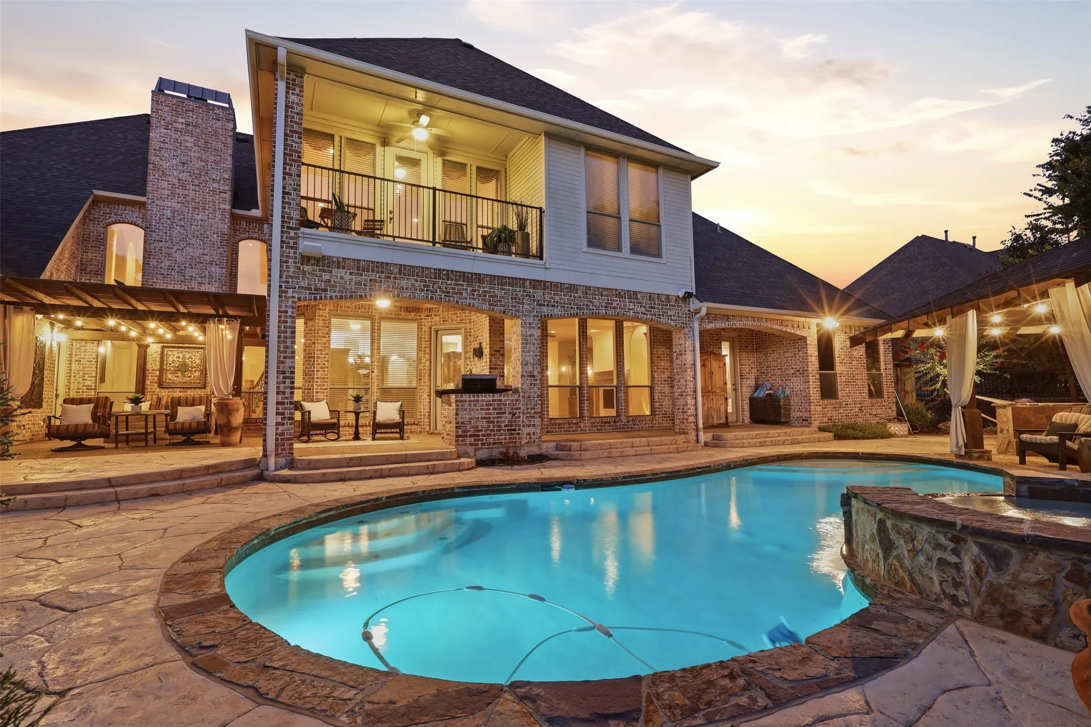 Rear exterior featuring a stone patio, built-in swimming pool, covered outdoor living area, and a second-story balcony