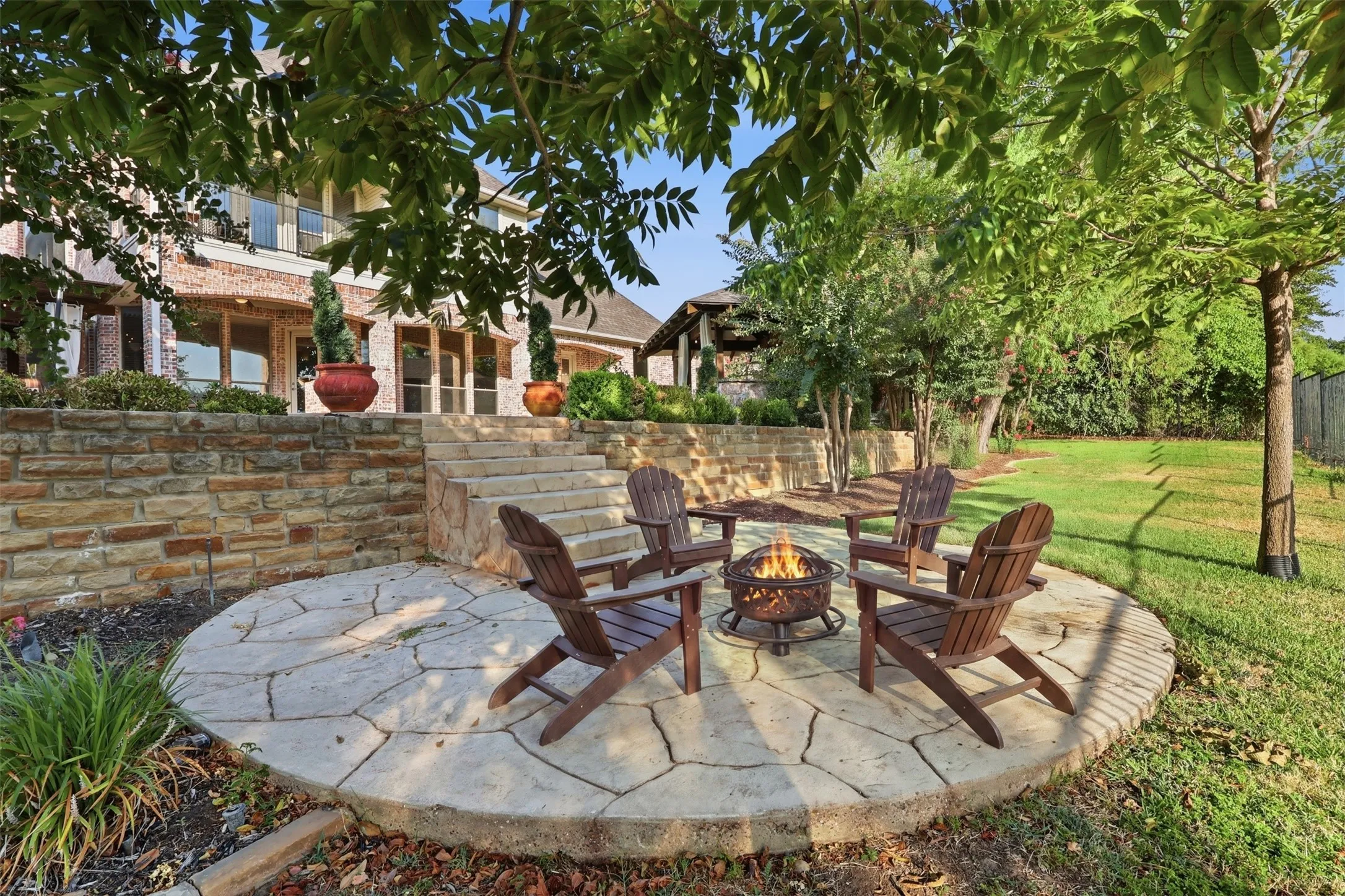 Stone-retained patio featuring a circular flagstone hardscape, multiple mature trees, and a tiered stone staircase leading to the residence
