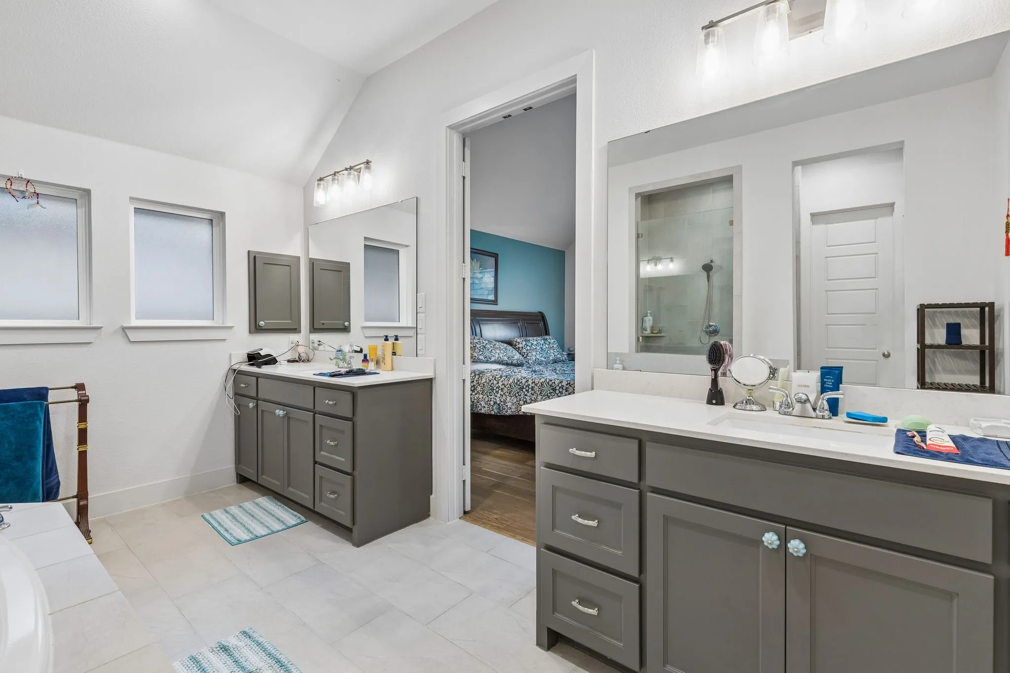Spacious bathroom featuring twin gray vanity cabinets with white countertops, light tile flooring, and two frosted windows