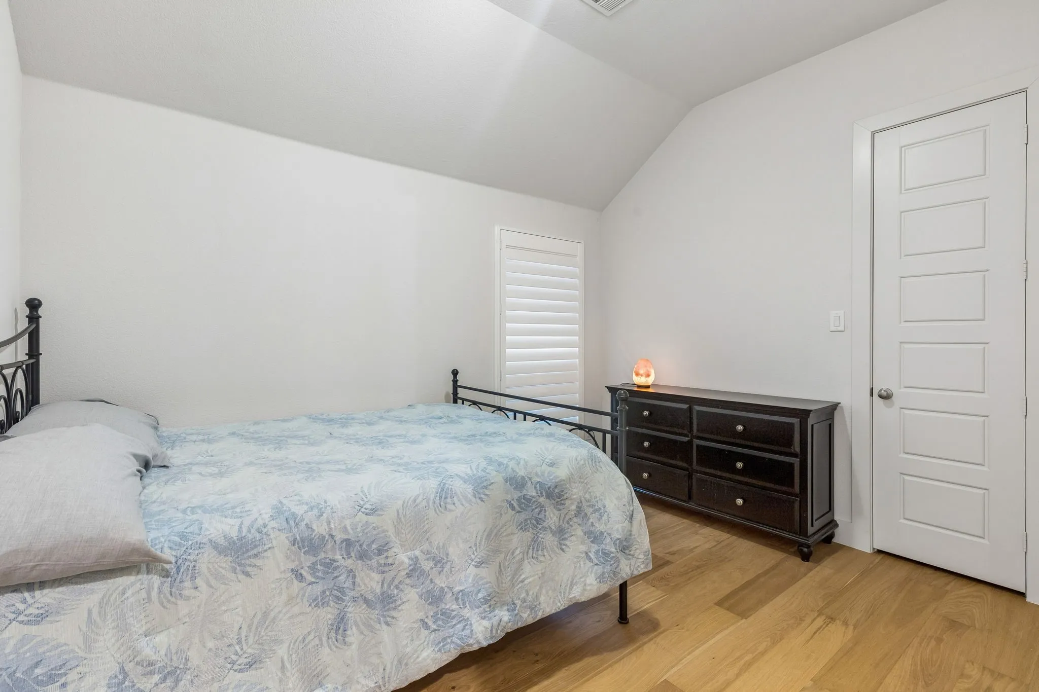 Interior room featuring light wood-finish flooring, a white five-panel interior door, and a window with white louvered shutters