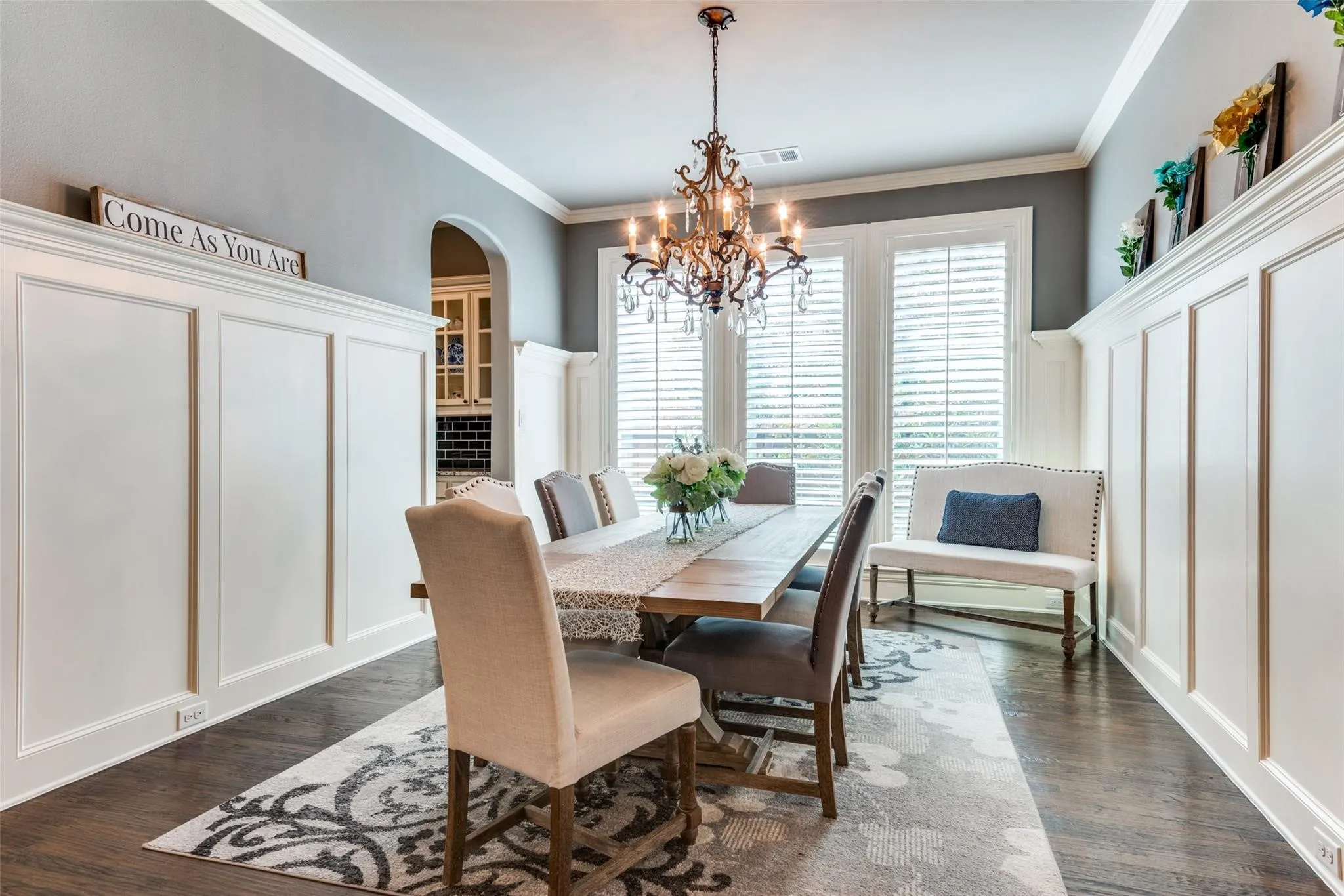 Designed to impress, this elegant formal dining room offers the perfect setting for memorable dinners and special occasions. Rich hardwood floors, sophisticated wainscoting, and extensive crown molding create a timeless backdrop, while the designer chandelier adds a touch of glamour and warmth. Tall windows dressed with plantation shutters fill the space with natural light, enhancing the room’s inviting ambiance.