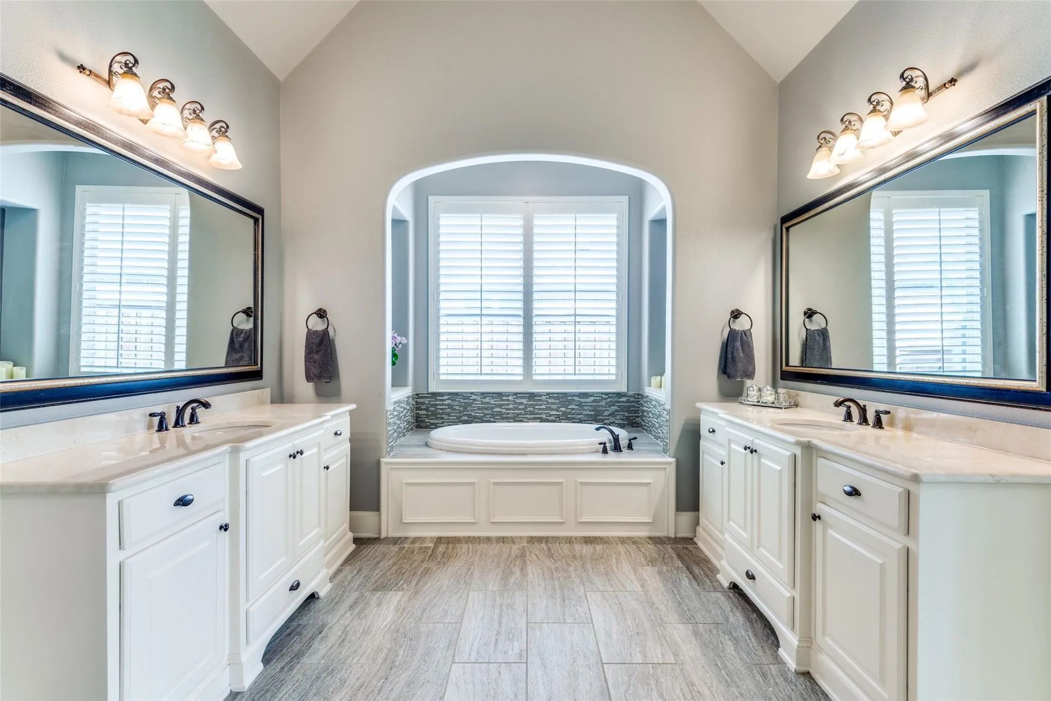 Step into a spa-inspired ensuite bath designed to elevate everyday living. This luxurious retreat showcases dual oversized vanities with abundant cabinet storage, elegant countertops, and expansive framed mirrors that create a bright and sophisticated atmosphere. At the center of the space, a relaxing soaking tub is beautifully nestled within an arched alcove, accented by designer tile surround and natural light streaming through plantation-shuttered windows.