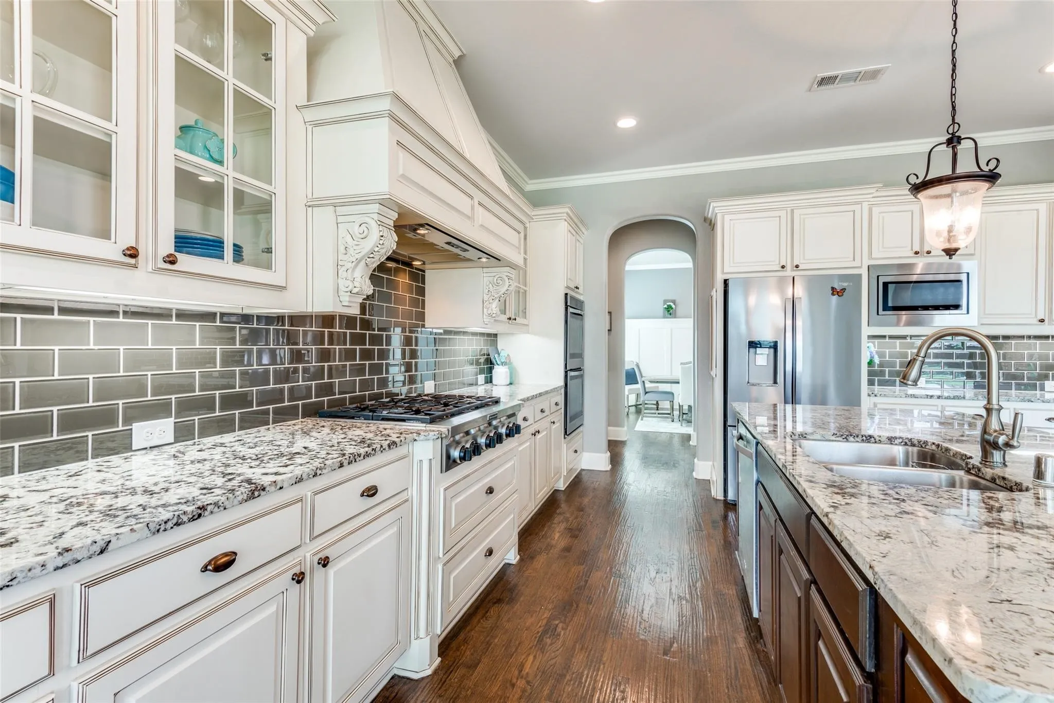 Rich granite countertops complement the custom cabinetry, while an abundance of cabinets—including glass-front display cabinets—provide exceptional storage and showcase space. The professional-grade gas cooktop is highlighted by a striking designer vent hood, creating a stunning focal point. Stainless steel appliances, double ovens, and an oversized pantry complete this impressive culinary space.
