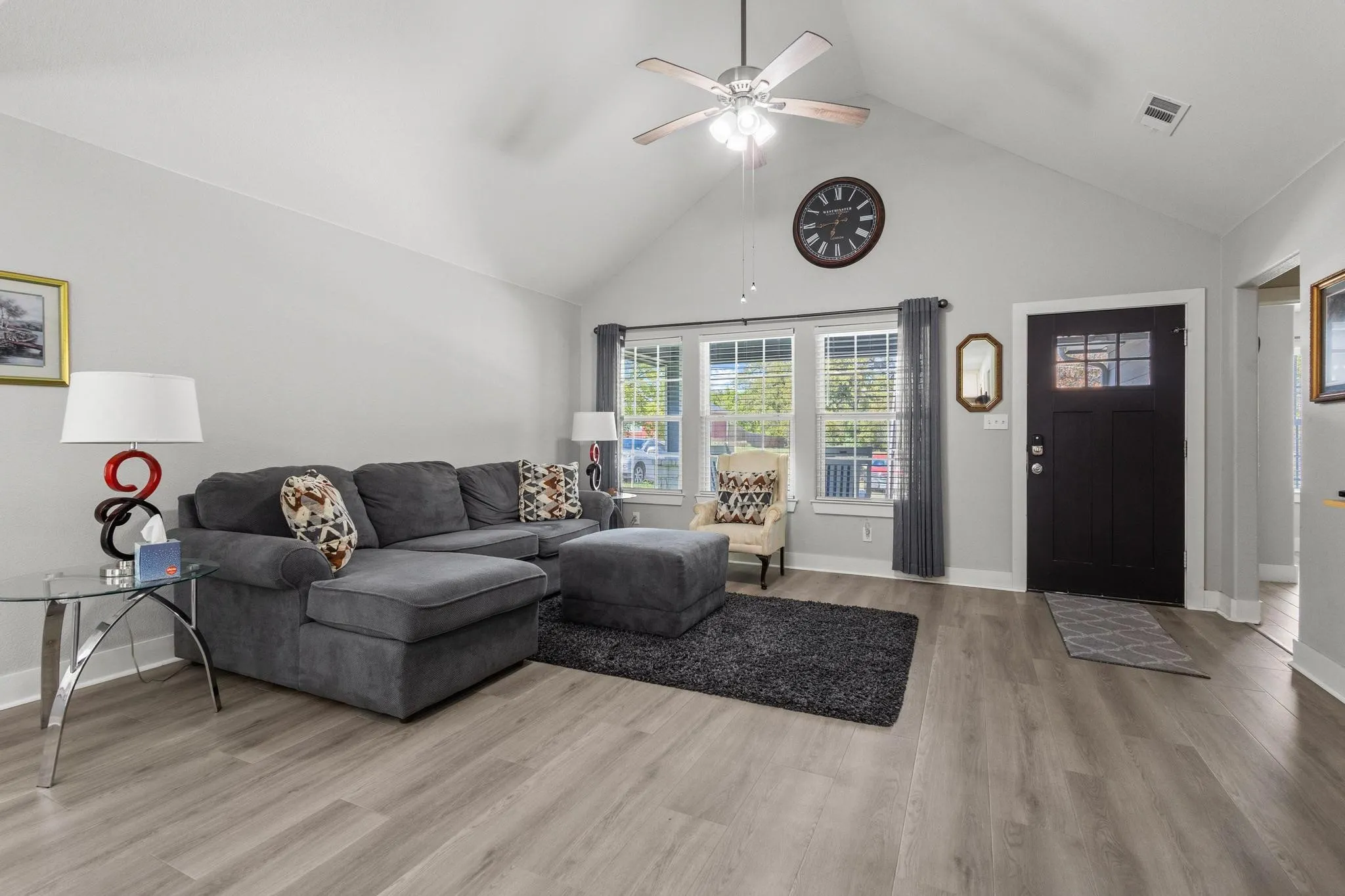Living area featuring a vaulted ceiling with a ceiling fan, light wood-finish flooring, a black front door with upper window panes, and three large windows with white blinds