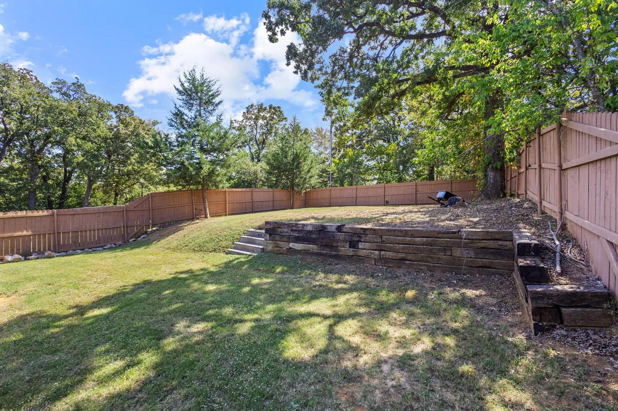 Expansive backyard featuring a tiered landscape, privacy fencing, mature trees, wood retaining walls, and a grassy lawn