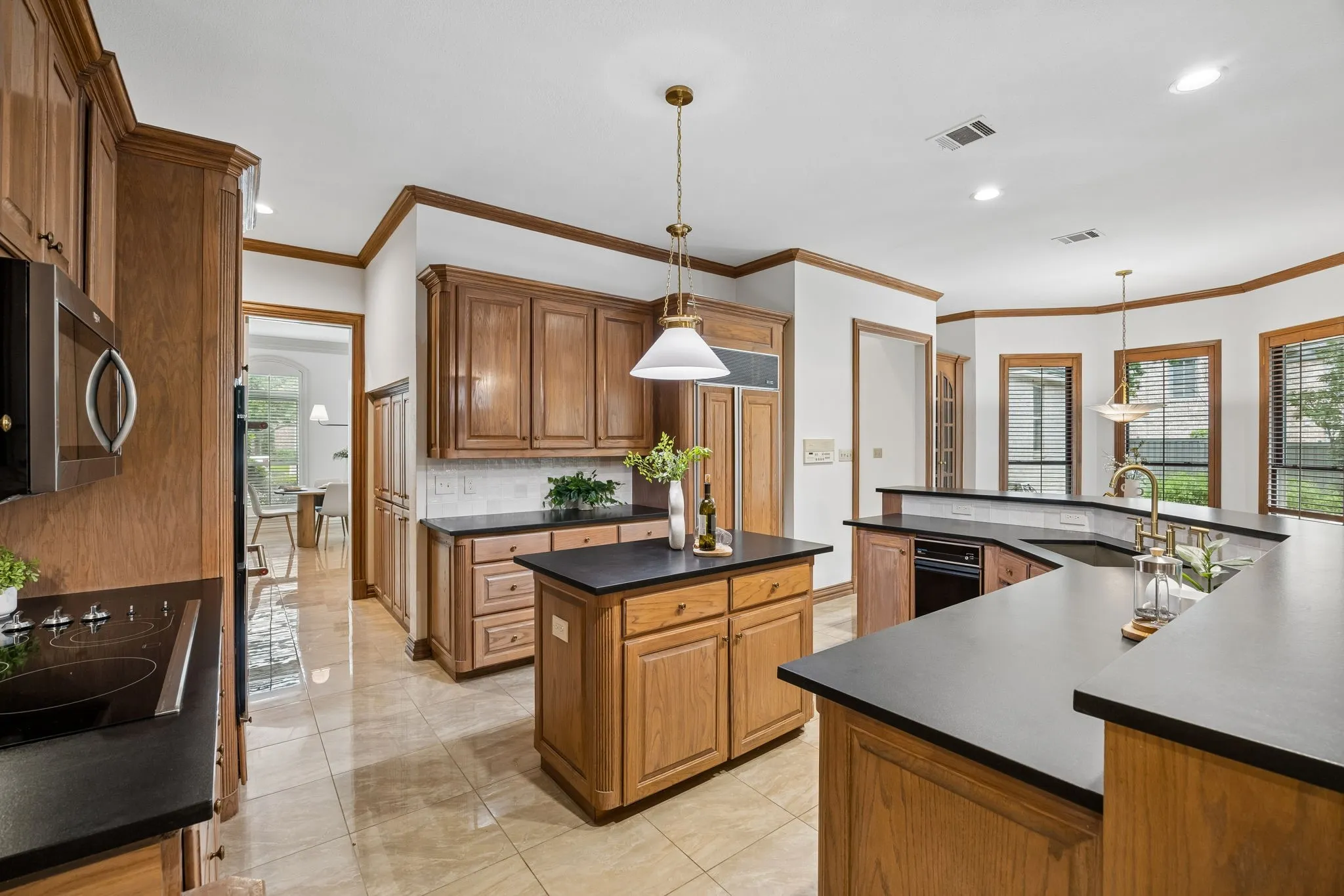 Spacious kitchen featuring extensive wood cabinetry, dark countertops, a central island with pendant lighting, integrated appliances, and light-toned tile flooring