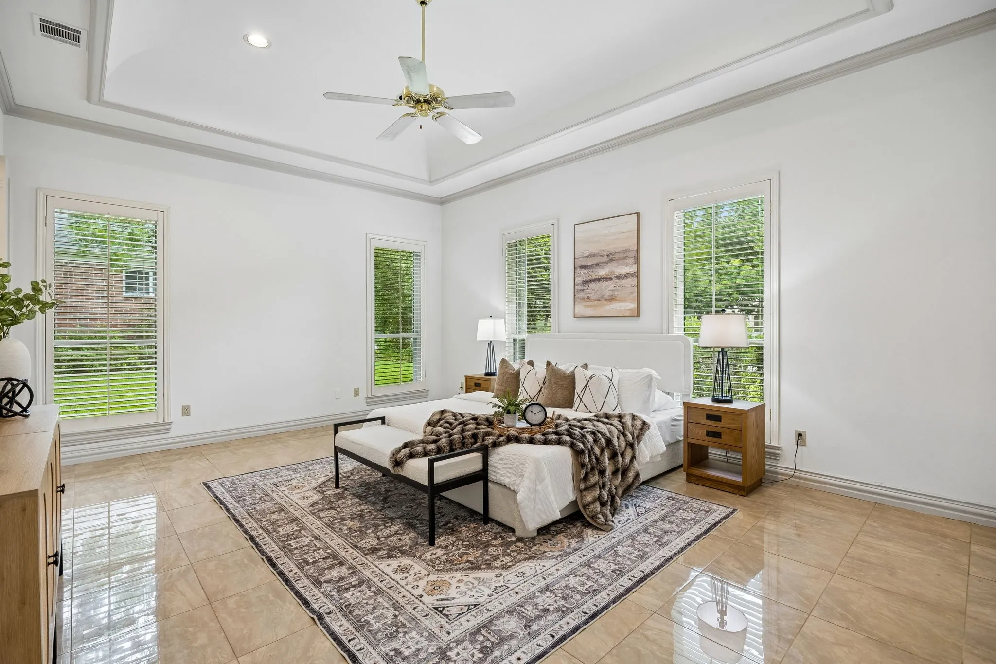 Spacious bedroom featuring light-toned tile flooring, a tray ceiling with a ceiling fan, and four large windows with blinds