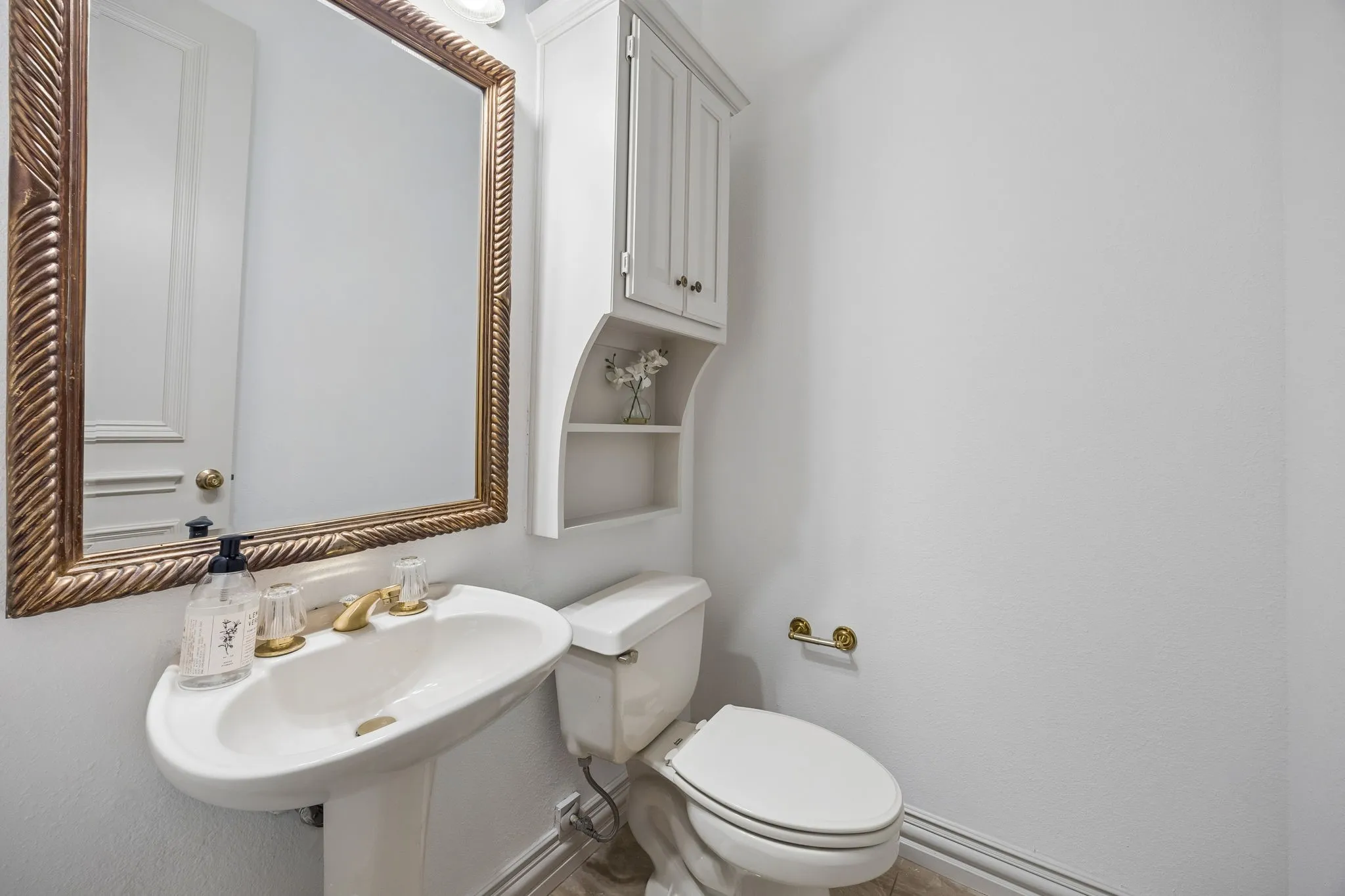 Pedestal sink with brass-finish faucet, framed mirror, built-in wall cabinet with open shelving, toilet, and brass-finish toilet paper holder