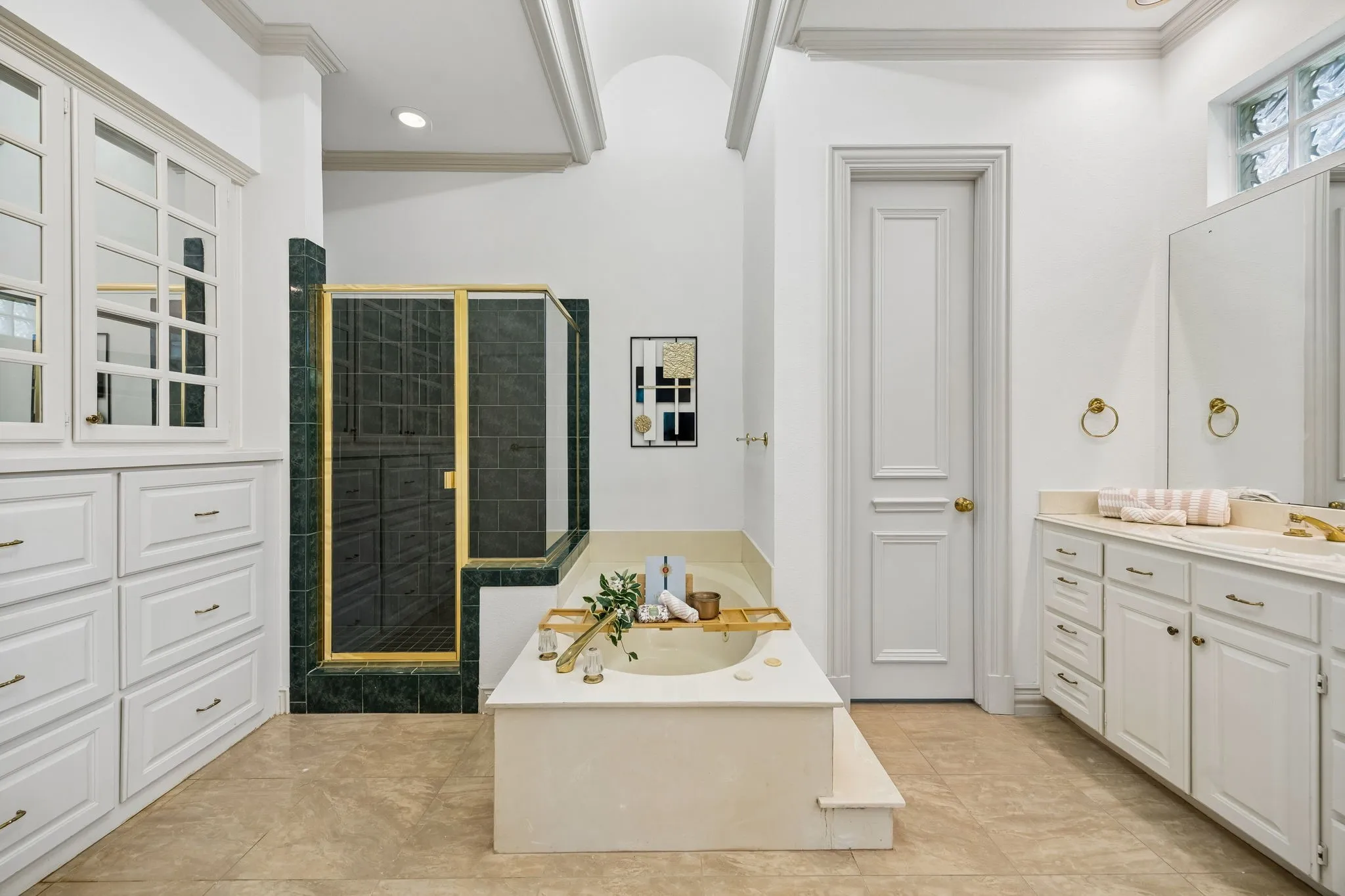 Bathroom featuring a built-in soaking tub with steps, a glass-enclosed shower with green and dark tile, ample white cabinetry with gold-toned hardware, a double vanity with an integrated sink, and light-toned tile flooring