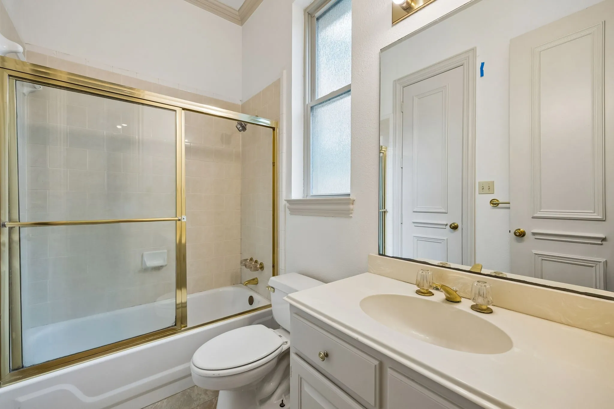 Bathroom featuring a tub/shower combination with gold-tone framed glass doors, a vanity with an integrated sink and gold-tone faucet, a large wall-mounted mirror, and a frosted window