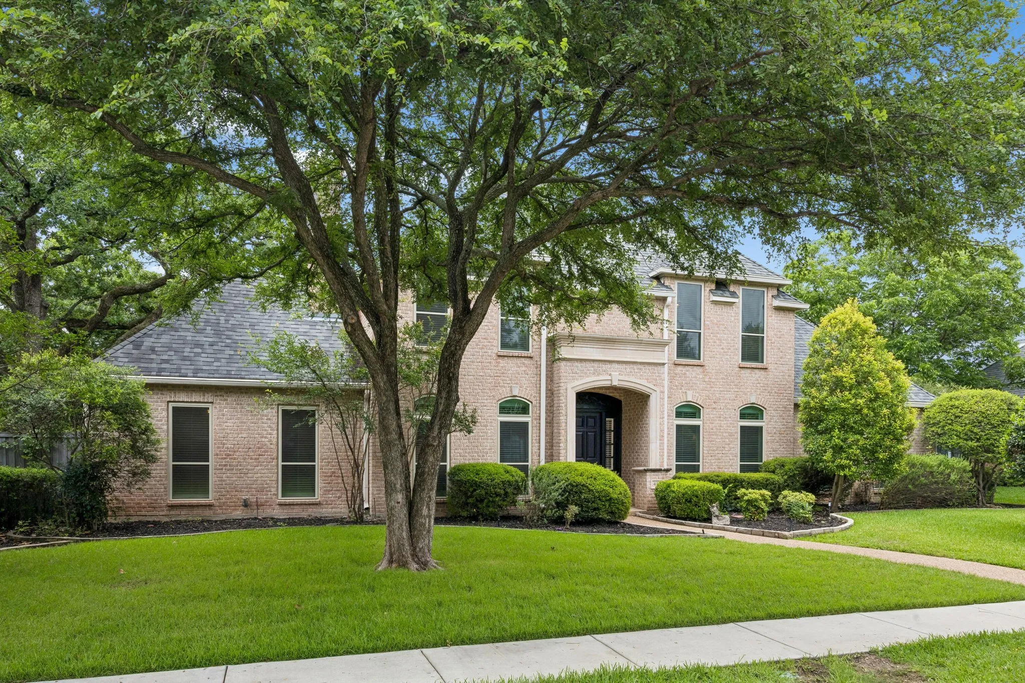 Two-story brick exterior with a prominent arched entry, multiple windows, a grey shingle roof, and a mature tree