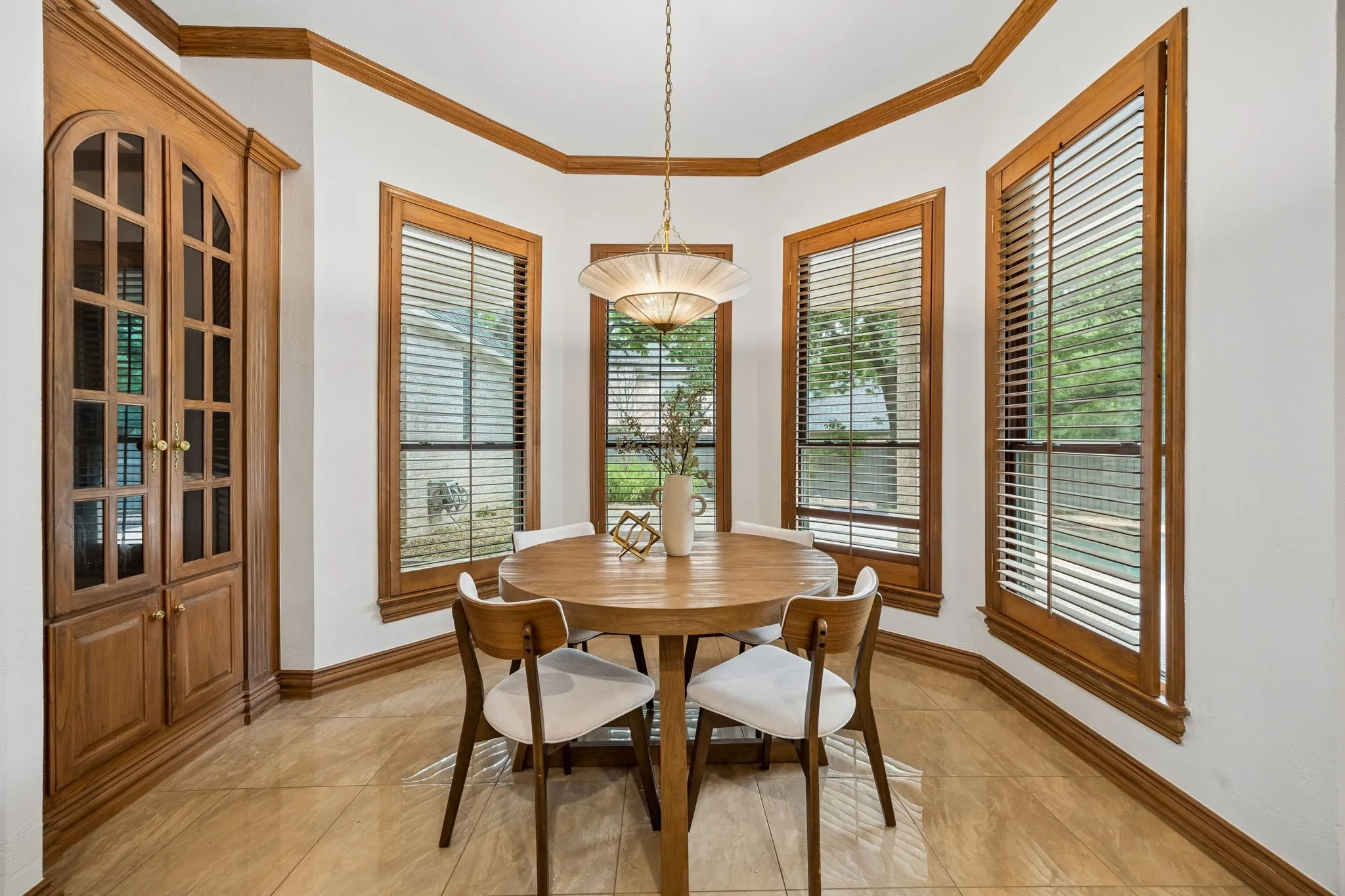 Bay window dining area featuring three windows with wood blinds and trim, a chandelier, a built-in wood-finish cabinet with glass panel doors, and tile flooring