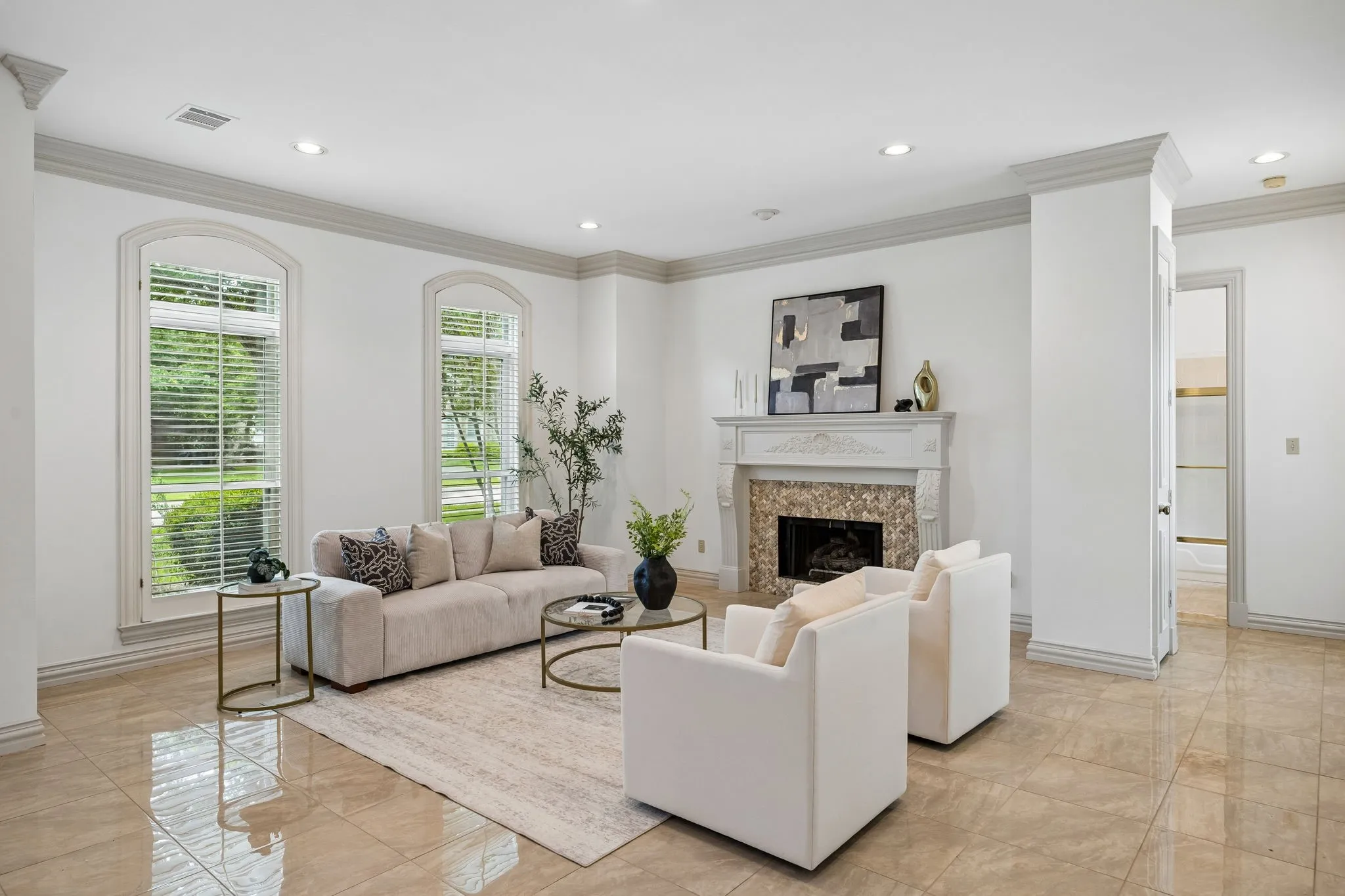 Spacious living area featuring polished tile flooring, a decorative stone fireplace with an ornate mantel, two arched windows with blinds, recessed ceiling lighting, and detailed crown molding throughout