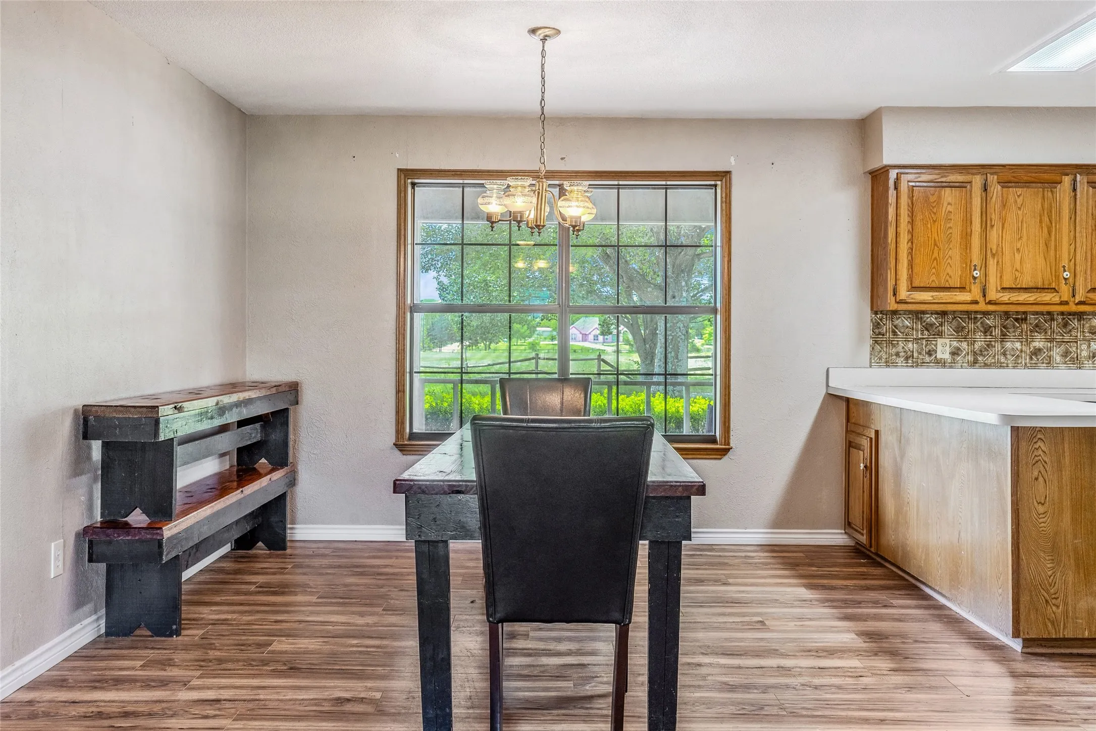 Dining area featuring wood-finish flooring, a window with a brass-finish chandelier, and a partial view of the kitchen with wood cabinetry and light-toned countertops