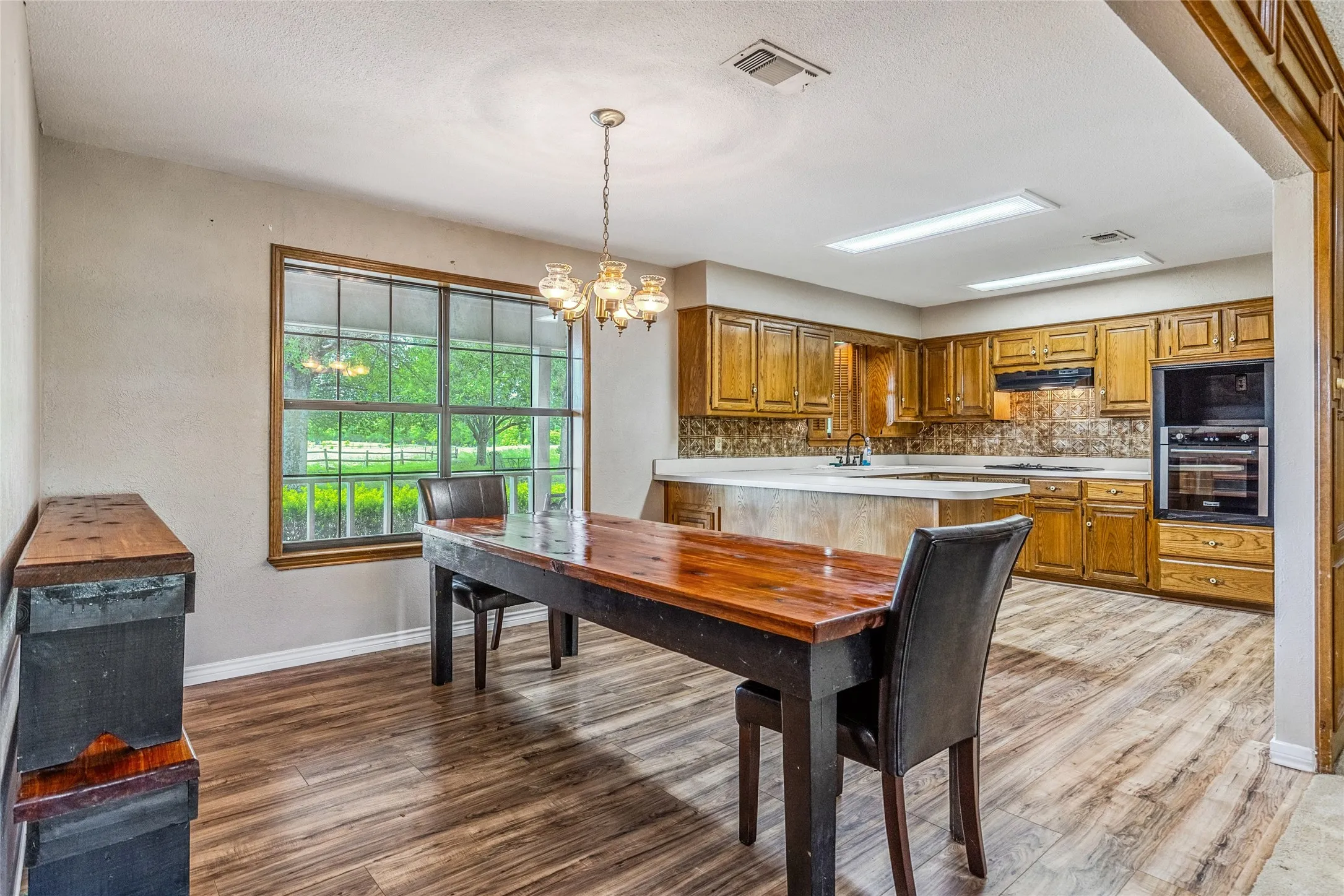 Open concept kitchen and dining area featuring wood-finish flooring, a large window, a chandelier, wood cabinetry, and an integrated double oven