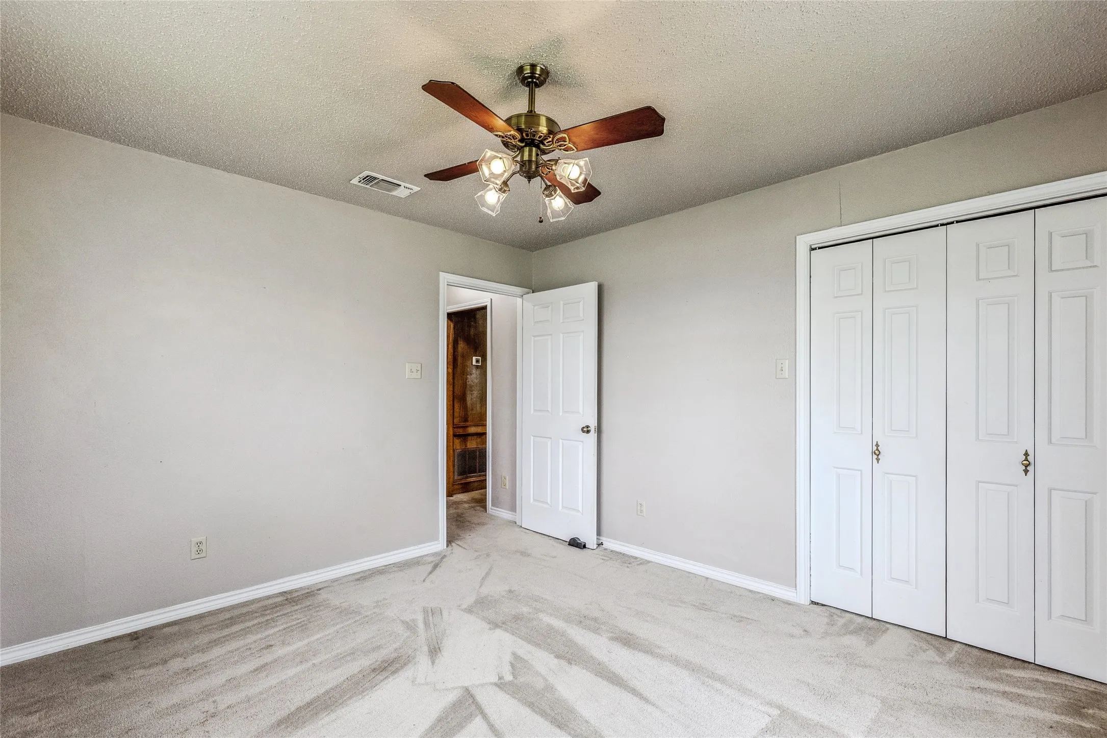 Room featuring light-toned walls, ceiling fan with wood-finish blades and brass-tone hardware, textured carpeting, and a white bifold closet