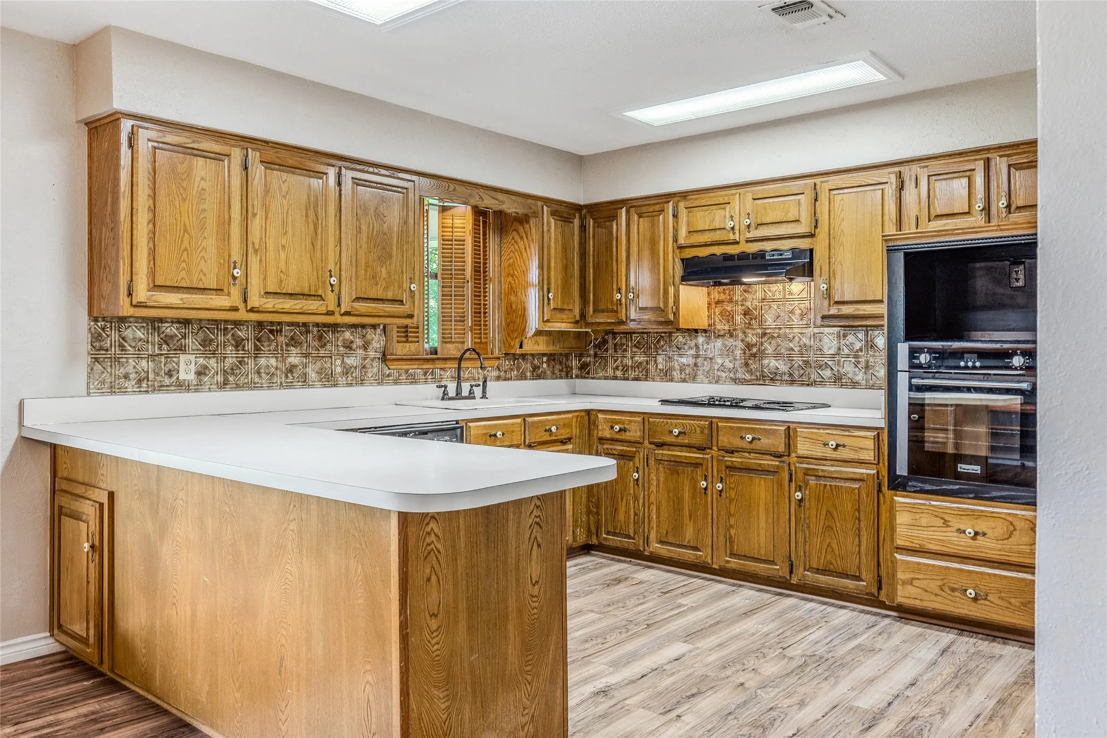 Kitchen featuring wood-finish cabinetry, white countertops, a built-in oven, integrated cooktop, and patterned backsplash