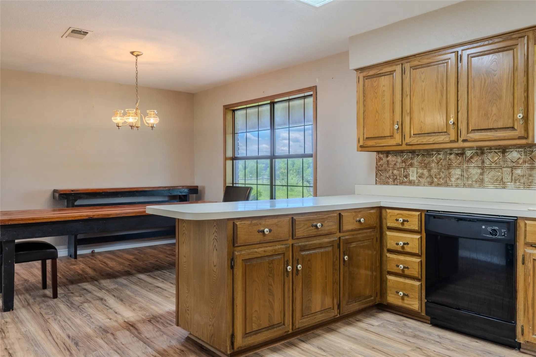 Kitchen space featuring wood-finish flooring, oak cabinetry, a light-toned countertop, a black built-in dishwasher, and a patterned backsplash