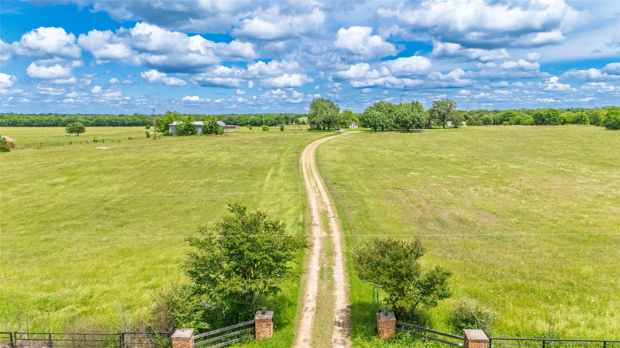 Expansive rural landscape featuring a winding gravel driveway, mature trees, open pastures, and a distant building