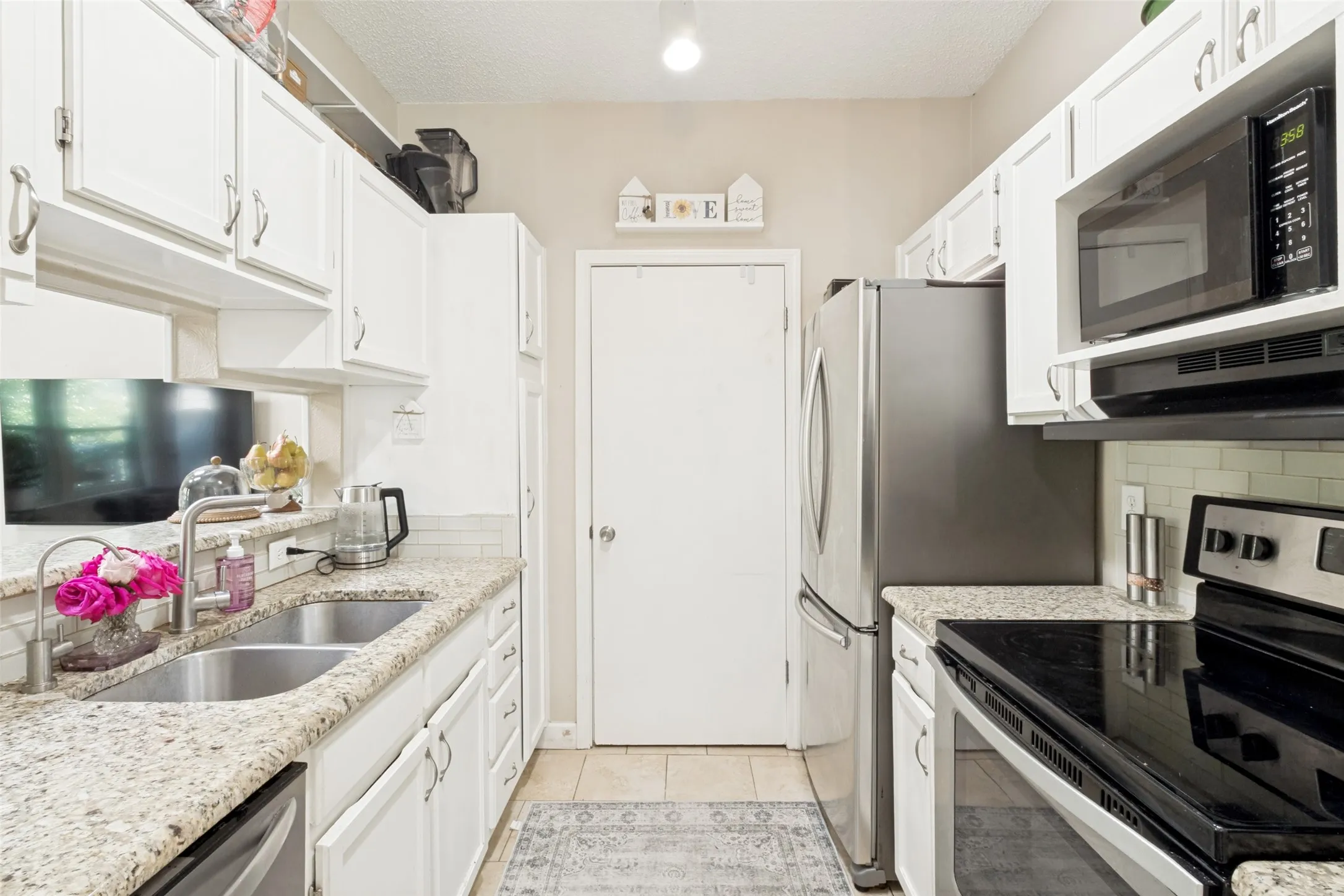Galley kitchen featuring white cabinetry, light-toned granite-style countertops, stainless steel appliances, a double basin sink, and tile flooring