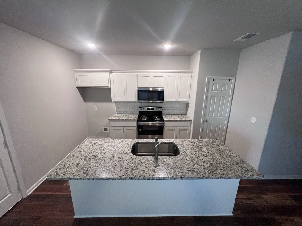 Kitchen featuring a central island with an undermount sink, granite-finish countertops, white shaker cabinetry, stainless steel appliances, and wood-finish flooring