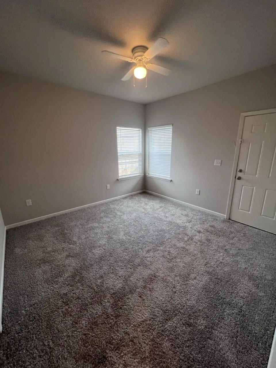 Carpeted room featuring neutral-toned walls, corner windows with blinds, a ceiling fan with light, and white trim